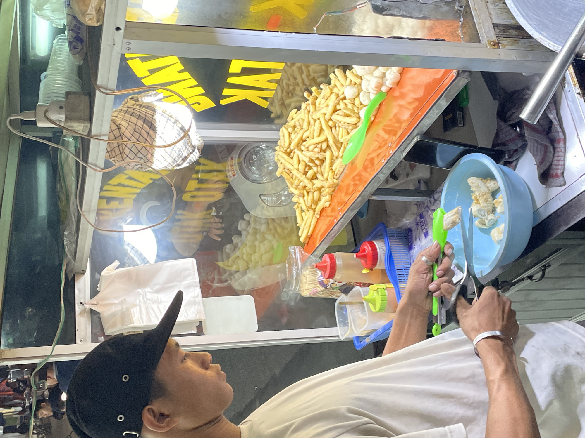 A person wearing a black baseball cap and a white t-shirt is seen from the chest up, looking upwards and to the left. They are at a food stall, with various food items and condiments visible. In the foreground, the person's hands are holding a pair of tongs and scissors, seemingly preparing food.
The food stall appears to be outdoors, possibly at night given the lighting. Behind the person, a display case holds a variety of fried snacks, including french fries and puffed snacks, arranged on an orange tray. There are also bottles of sauces and condiments, such as ketchup and chili sauce, in plastic dispensers. A blue basket holds some food items, and a light blue bowl contains more prepared food.
White plastic bags are stacked on a shelf behind the stall. Visible text on the stall's signage appears to be in yellow capital letters, with "LVK" being partially decipherable. The overall scene suggests a busy street food vendor preparing orders for customers in North Jakarta, Indonesia. The person's gaze is directed upwards, and their facial expression is neutral, suggesting a focus on their task.