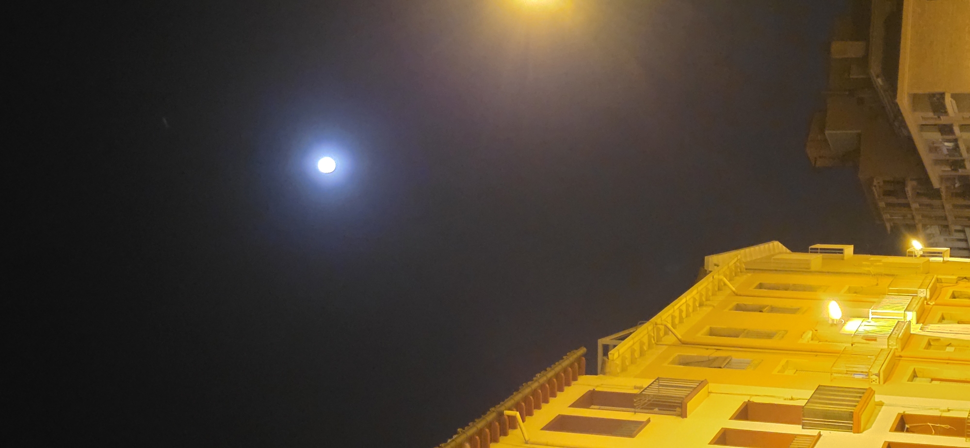 A low-angle photograph taken at night in Huesca, Spain, shows a portion of a building in the foreground, and the sky with a moon-like object in the background. The building is illuminated with yellow-orange light, and its windows and architectural details are visible. The building appears to be tall, extending upwards. In the sky, a bright circular object, resembling the moon, is visible with a faint aura around it. A streetlamp is also emitting light near the top of the frame. The sky appears dark.