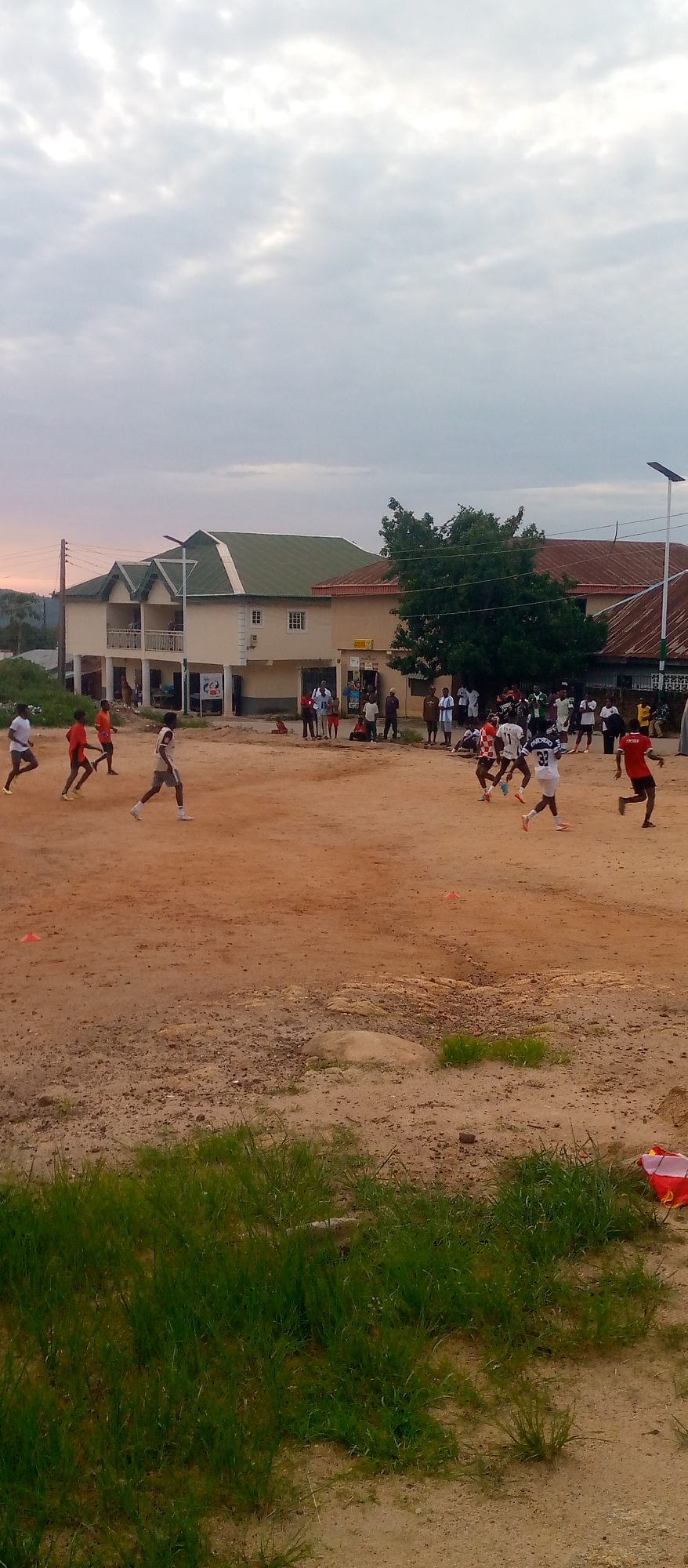 The image depicts an outdoor scene on a dusty field, likely a sports ground, where a group of people are engaged in a soccer game. The sky above is overcast with diffused light, suggesting it might be early morning or late afternoon. In the background, there are several buildings with varied architectural styles, including a light-colored structure with a green roof and balconies, and another building with a corrugated metal roof. A solar-powered light pole stands near the buildings.
Several individuals are visible on the field, actively participating in or observing the soccer match. Some players are in motion, kicking or running with the ball. The players are wearing athletic attire, with some in jerseys and shorts. In the foreground, there is overgrown grass and some scattered debris. The ground is predominantly reddish-brown earth, indicative of a dry, possibly arid environment. The overall atmosphere suggests a casual sporting event taking place in a community setting.