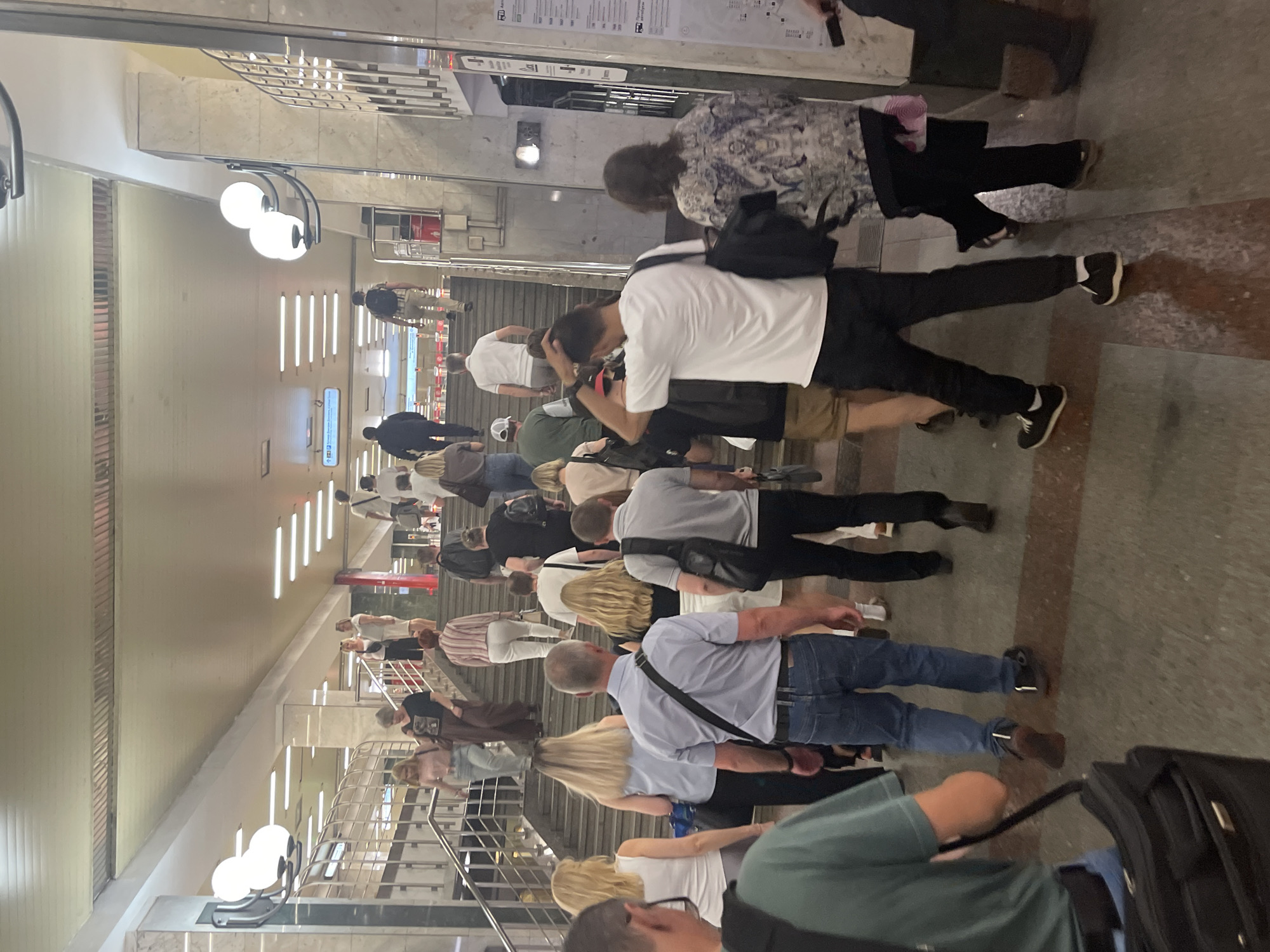The image depicts a group of people in an indoor, likely public, setting. The scene appears to be a subway or metro station in Moscow, Russia, suggested by the presence of escalators, signage that resembles Cyrillic script, and the overall architecture. People are seen walking on stairs and escalators, some carrying bags and backpacks. The lighting suggests it is either daytime or well-lit indoor lighting. There's no indication of specific weather conditions as the scene is indoors. The people are dressed casually, and their expressions are not clearly discernible due to the distance and angle. One sign appears to have text, possibly directions or station information, but it is illegible. The overall atmosphere seems to be one of transit and everyday movement.