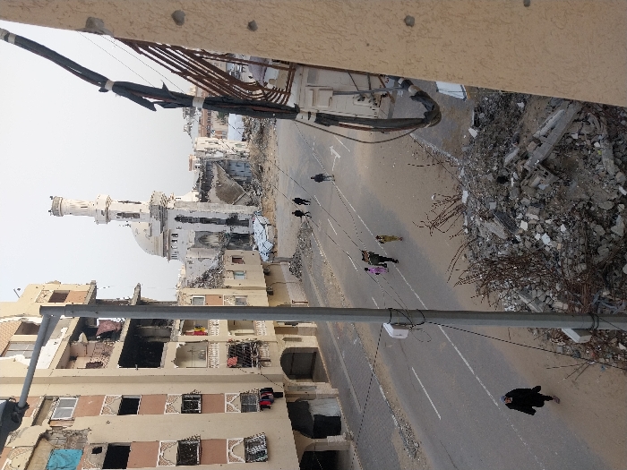 An elevated view captures a street scene in Khan Yunis, Palestinian Territory, characterized by extensive structural damage. The central feature is a heavily damaged mosque with a white dome and a tall, partially intact minaret, positioned at the end of the visible street. Surrounding the mosque and lining the street are multi-story buildings, all displaying significant destruction, including blown-out windows, missing wall sections, exposed interiors, and visible rebar.
On the left side, a large apartment building exhibits numerous broken windows, damaged balconies, and dark openings where parts of the structure are missing. Laundry is visible hanging from some windows or balconies. On the far right, a beige building wall in the foreground shows at least two distinct bullet or shrapnel holes, with a bundle of thick, damaged cables hanging down.
The asphalt road running through the center is largely clear but flanked by widespread piles of concrete rubble, twisted metal rebar, and debris. A prominent grey utility pole stands in the left foreground, supporting overhead wires that span across the street.
Several individuals are present on the street. In the middle ground, a group of four people walk together; one appears to be carrying an item over their shoulder, while another, possibly a child, is walking closely with an adult. Further down the street, two individuals in dark clothing walk side-by-side towards the mosque. In the immediate foreground on the left, a single person dressed in dark clothing walks on the road's shoulder. Another lone figure is visible walking on the far right side of the street, closer to the mosque. The sky is a uniform, light grey, indicating an overcast or hazy day.