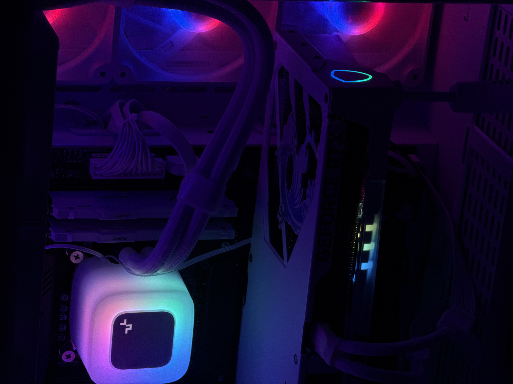 The image depicts the interior of a white computer tower case illuminated by colorful RGB lighting. A white CPU cooler with a glowing, multifaceted light effect is visible in the upper left corner, displaying shades of blue, green, purple, and pink. To its right are several white RAM modules, some labeled "FURY." A white GPU with the "GEFORCE RTX" logo visible is mounted horizontally in the lower half of the frame. This GPU also features RGB lighting, with a blue and green glowing outline visible on its side. Two large, white computer fans are situated on the right side of the case, emitting pink and blue light. The overall environment is dark, with the RGB lighting being the primary light source, creating a vibrant and high-tech aesthetic. There are no people or any indications of the city of Poritsy, Russia within the image.