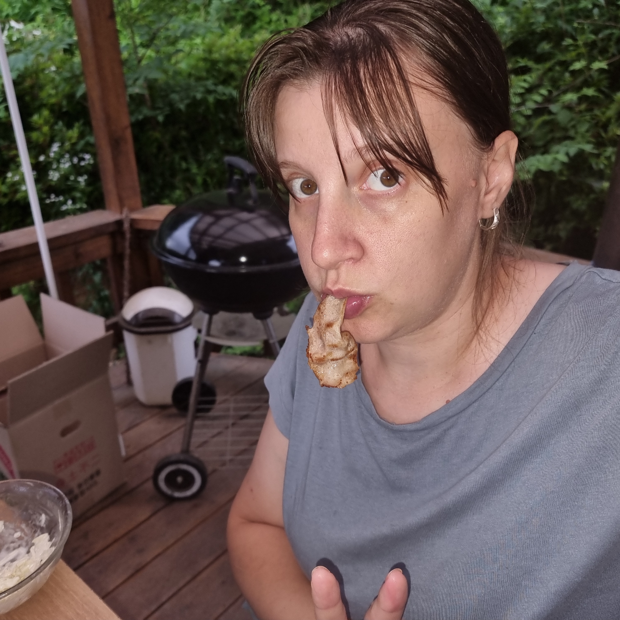 A woman is shown on a wooden deck, possibly a porch or patio. She is holding a piece of food, perhaps bacon, to her mouth. She has brown hair and is wearing a grey t-shirt. In the background, a black grill with a lid is visible, along with a white trash can. There are also some cardboard boxes on the deck. The overall setting appears to be outdoors, with greenery and trees visible in the background, suggesting a natural or semi-rural environment. The lighting suggests it is daytime, possibly overcast or shaded. The woman's expression could be interpreted as playful or surprised, as she makes eye contact with the camera while holding the food.