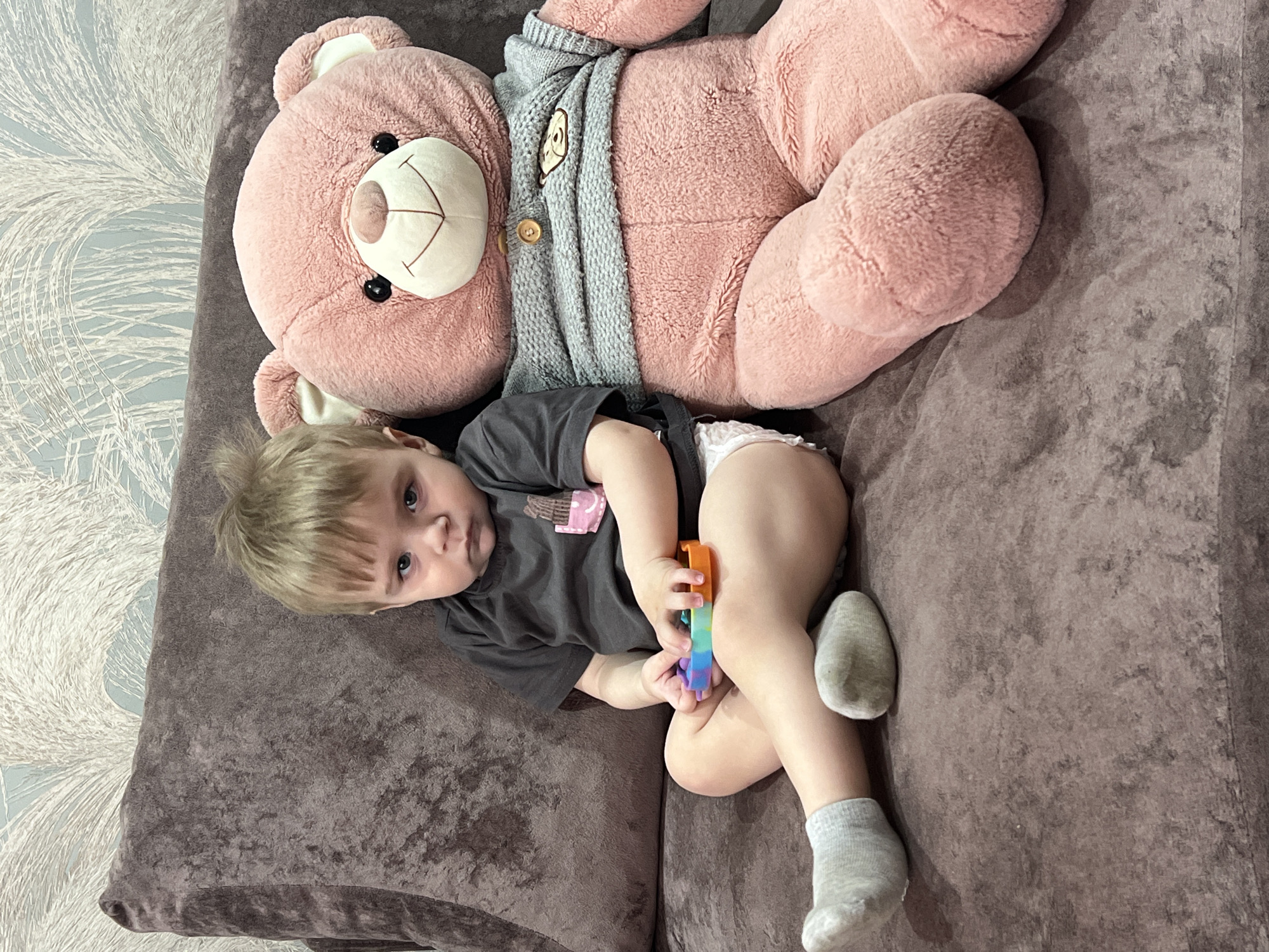 A young child and a large pink teddy bear are depicted lying on a dark brown plush sofa in an indoor setting in Noviy, Russia. The child, a toddler with light blonde hair, is wearing a dark grey t-shirt featuring a graphic of a pink and brown multi-layered dessert, a white diaper, and light grey socks. They are holding a rainbow-colored pop-it fidget toy. The child is looking directly at the camera with a neutral expression.
The oversized pink teddy bear, dressed in a grey knitted sweater with a small bear face patch, is positioned behind the child, resting on the sofa. To the left of the sofa, wallpaper with a light blue background and a prominent white, feathery or leafy pattern is visible. The overall scene suggests a calm, domestic moment during the daytime.