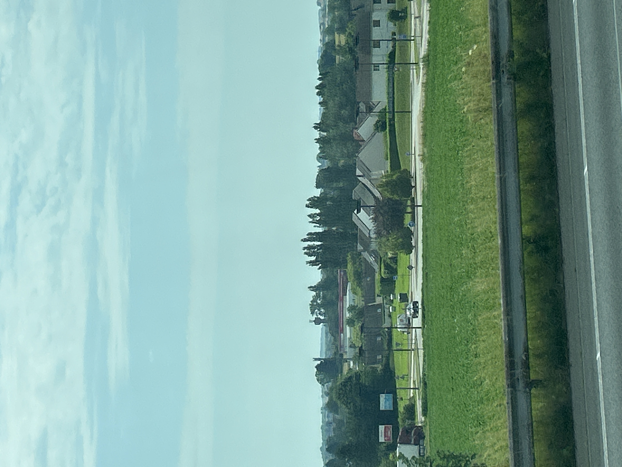 The image depicts a suburban or semi-rural landscape with a road on the right side and a field of green grass adjacent to it. Beyond the grass, there are some buildings and trees. The sky is visible in the upper left portion of the image and appears to be partly cloudy. The scene is captured from a slightly elevated perspective, possibly from a vehicle or a building.
The buildings appear to be residential or commercial, with some featuring pitched roofs. There are also several trees scattered throughout the landscape, some of which are quite tall. The road appears to be a multi-lane highway or a significant road, with visible lane markings.
The time of day appears to be daytime, with the sky suggesting either morning or afternoon. The weather seems to be clear with some clouds.
No people are clearly visible in the image. There are some vehicles on the road, including what appears to be a white car.
There are some signs visible in the distance, but the text is too blurry to read. The overall impression is one of a typical daytime scene in a developed area, likely in France given the location context provided. The scene is calm and ordinary, with no particular event or activity that stands out, other than the presence of the road and the surrounding environment.