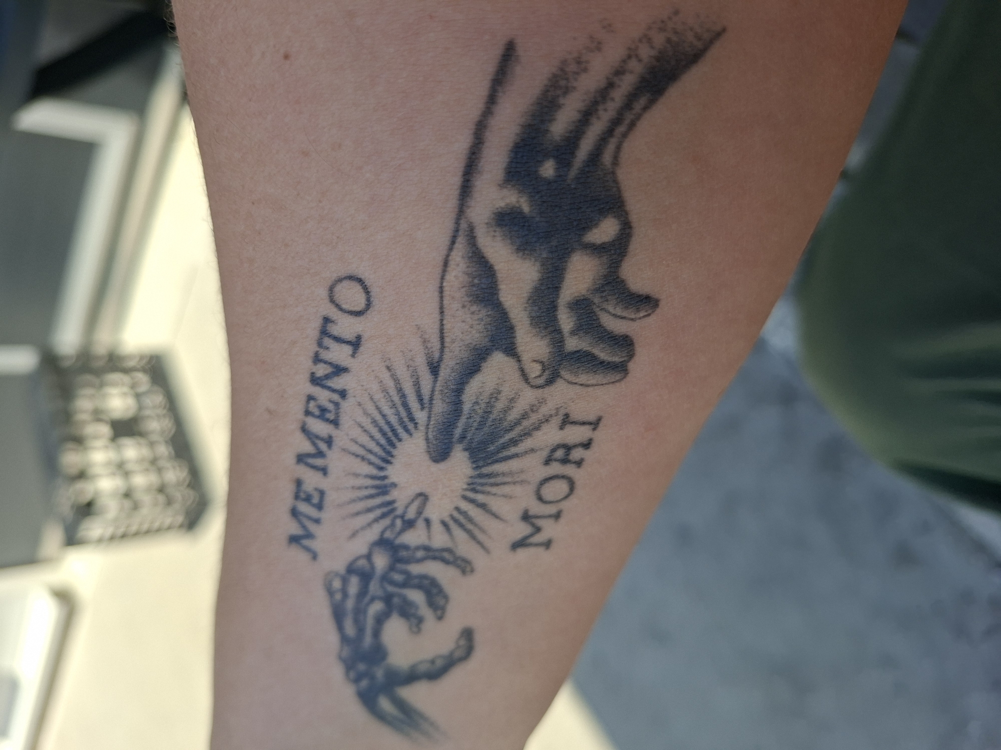 An arm has a tattoo featuring two hands reaching for each other with the text "Memento Mori" in a faded black ink. One hand appears to be a healthy, fleshy hand, while the other is a skeletal hand. A starburst is in between them. The arm is positioned in front of a blurry background that includes a white piece of furniture and what appears to be the corner of a green object of some sort. The arm is light-skinned.
There is no indication of Plovdiv, Bulgaria in the image.