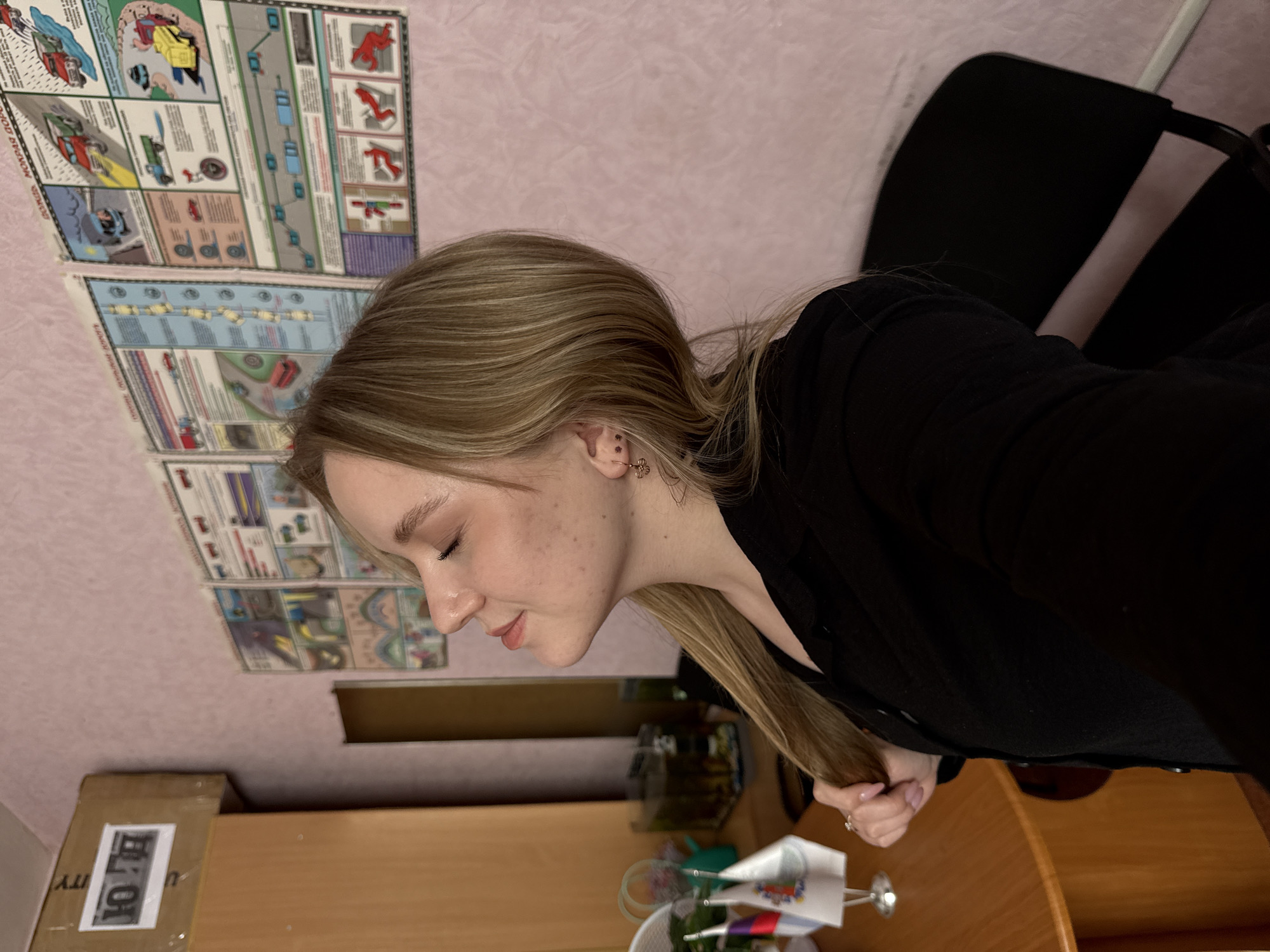 The image, rotated 90 degrees clockwise from its intended orientation, features a young woman in profile, likely in an indoor setting such as a study or home office in Chaikovsky, Russia. She has long, straight blonde hair and is wearing a dark, possibly black, long-sleeved top. Her eyes are gently closed, and a subtle, peaceful smile plays on her lips, suggesting a moment of calm or reflection. Her fair skin shows a scattering of freckles on her cheek, and she sports a small, golden, flower-shaped earring. Her left hand, adorned with light pink nail polish and a ring, is visible, gently holding a section of her hair.
The background consists of a light pink-textured wall covered with several large, multi-panel educational charts or posters. These posters prominently feature illustrations of cars, roads, and human figures, depicting various traffic rules and road safety scenarios. Partially visible text on one poster includes "ДОРОЖНАЯ..." (Road...) and "...ПОРЯ..." (Order/Rules...), while another clearly displays "ОПАСНОСТЬ!" (DANGER!) in red.
In the foreground and to the woman's left, a wooden desk or table is present. On the desk, a cardboard box bears a white label with partially discernible Russian letters "ТО" and "Ю". Nearby are a small fish tank or terrarium, a white basket, and a stand holding several small decorative flags, some displaying red, white, and blue colors, consistent with the Russian national flag. A portion of a dark office chair is also visible behind the woman. The lighting suggests an indoor setting, possibly daytime, with no direct outdoor light source apparent.