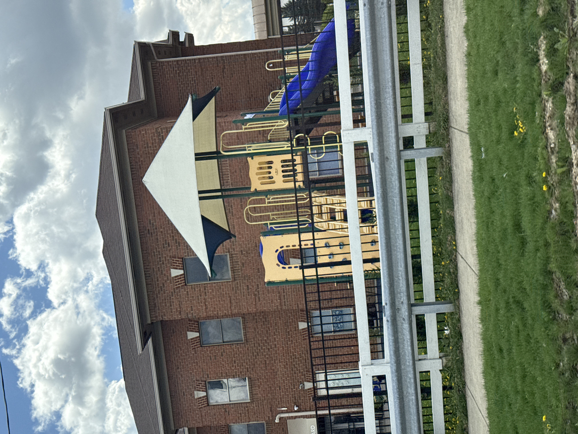 A medium-angle, slightly tilted, outdoor shot shows a playground and a brick building adjacent to each other. The playground equipment, featuring a blue slide and yellow and green structures, is partially obscured by a black metal fence and a white metal guardrail in the foreground. A three-story brick building is directly behind the playground. The building has multiple windows with dark frames, and a triangular shade structure is attached to the building, partially covering a portion of the playground. The sky, visible on the left side of the frame, is filled with white clouds. Grass and a sidewalk run along the right edge of the photo.
Without further context, pinpointing the exact location in Columbus, Ohio, is not possible.