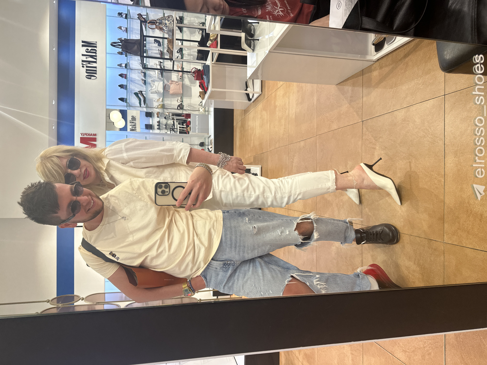 In Moscow, Russia, a couple is taking a mirrored selfie inside a shoe store. The man, on the left, wears ripped light-wash jeans, a cream t-shirt, sunglasses, and black sneakers. He is holding a phone to take the picture and has a large orange tote bag slung over his shoulder. The woman, on the right, wears white pants, a white collared shirt, and white mule heels. She is also wearing sunglasses and a large bracelet. The store's interior is visible in the background, with shelves displaying various shoes and handbags. The floor is tiled with large, light brown tiles. The store's branding, "MakFine," is visible on the wall.