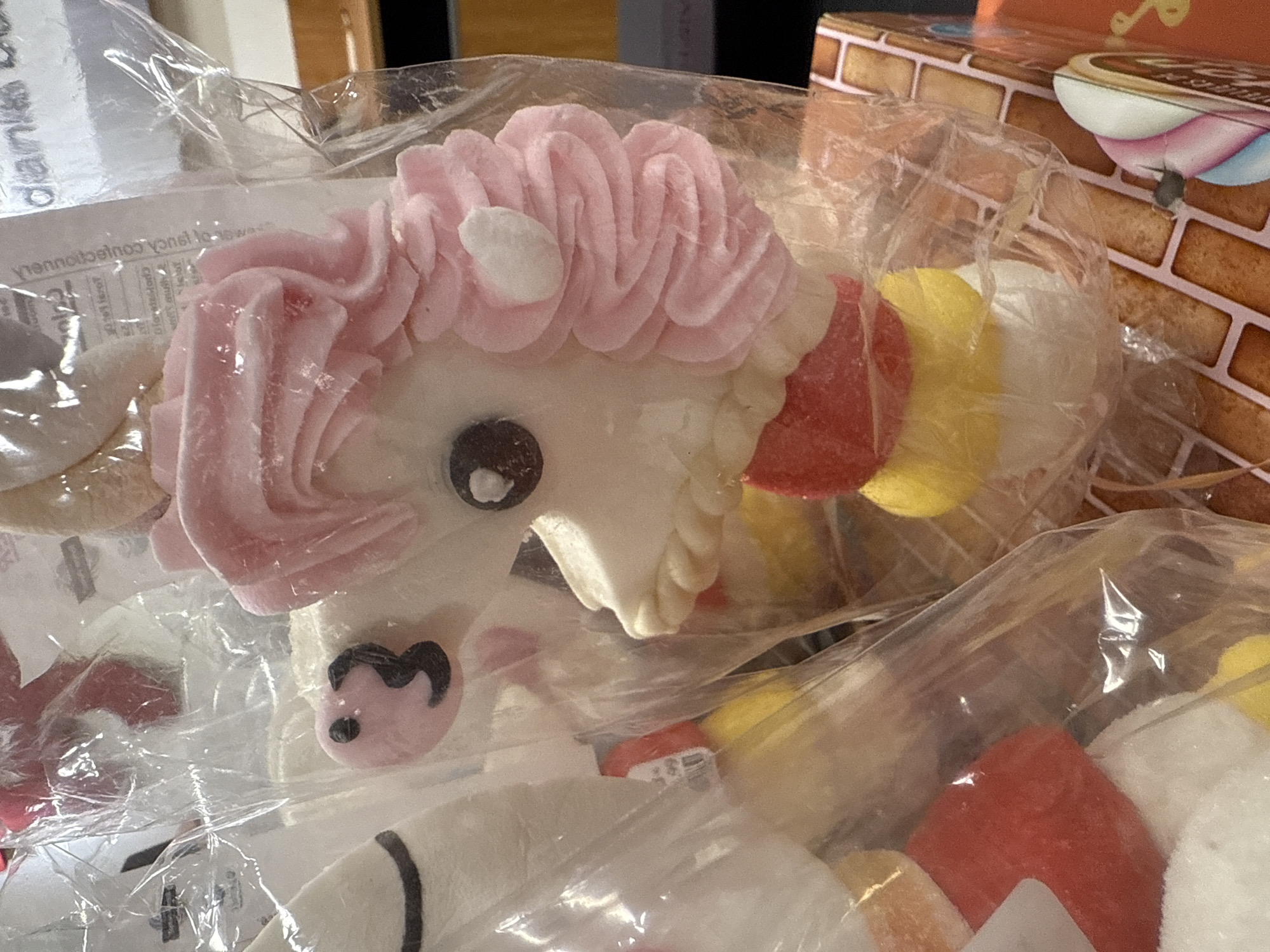 The image shows a close-up of packaged marshmallow candies shaped like unicorns and other figures. The candies are wrapped in clear plastic, suggesting they are commercially produced. A box with a brick-like design is visible in the background. The location is suggested to be Dania Beach, United States of America, though the image itself does not provide specific locational cues.