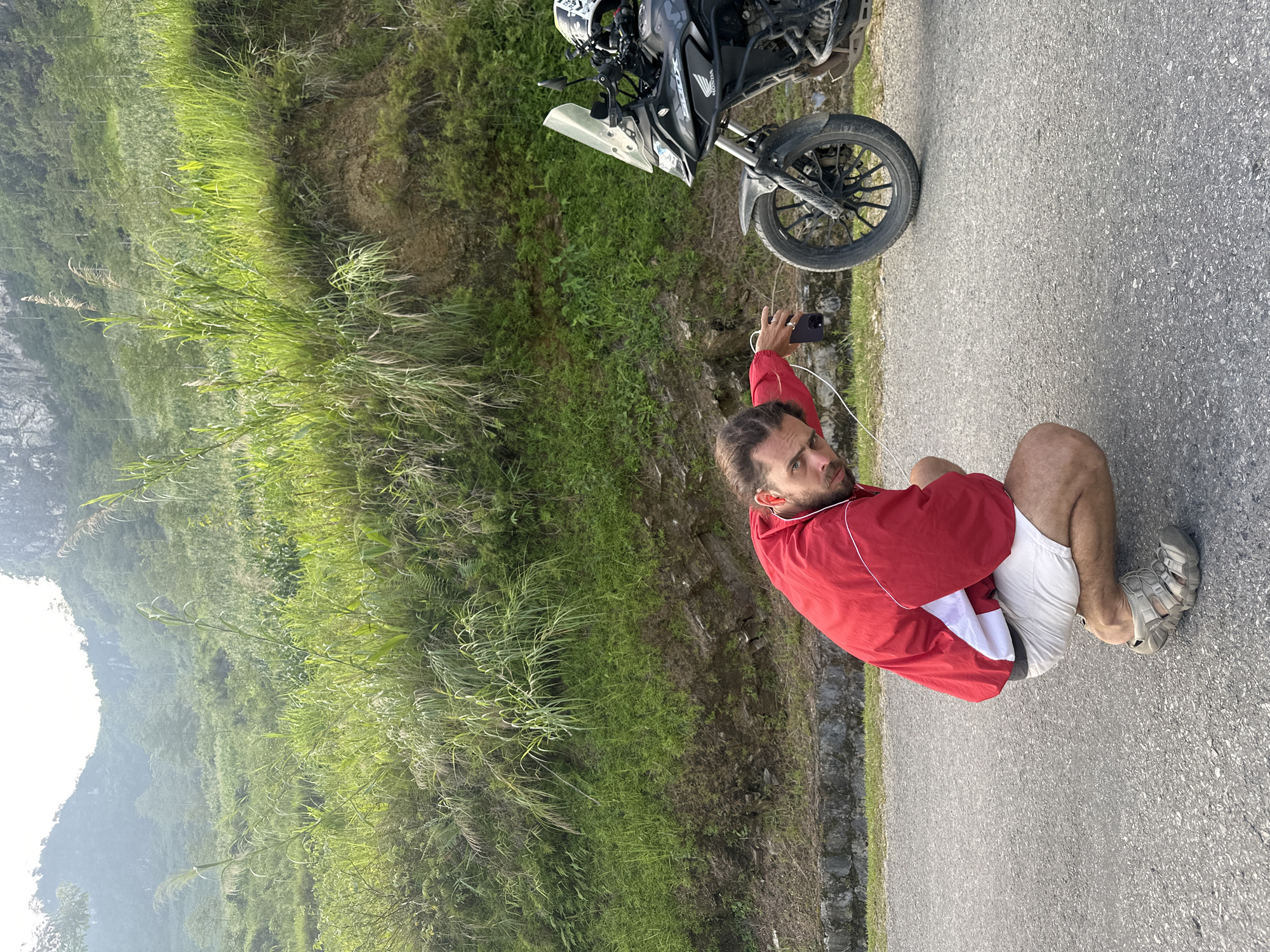 A man squats on an asphalt road alongside a parked motorcycle, holding a phone in his right hand and looking towards the bike. The motorcycle, with a partially visible "Honda" label and a helmet on top, is positioned on the right side of the road. The man is wearing a red jacket with white stripes, light-colored shorts, and sandals. Lush green vegetation covers the terrain adjacent to the road, and hills are visible in the background. The location is Du Già Commune, Vietnam.