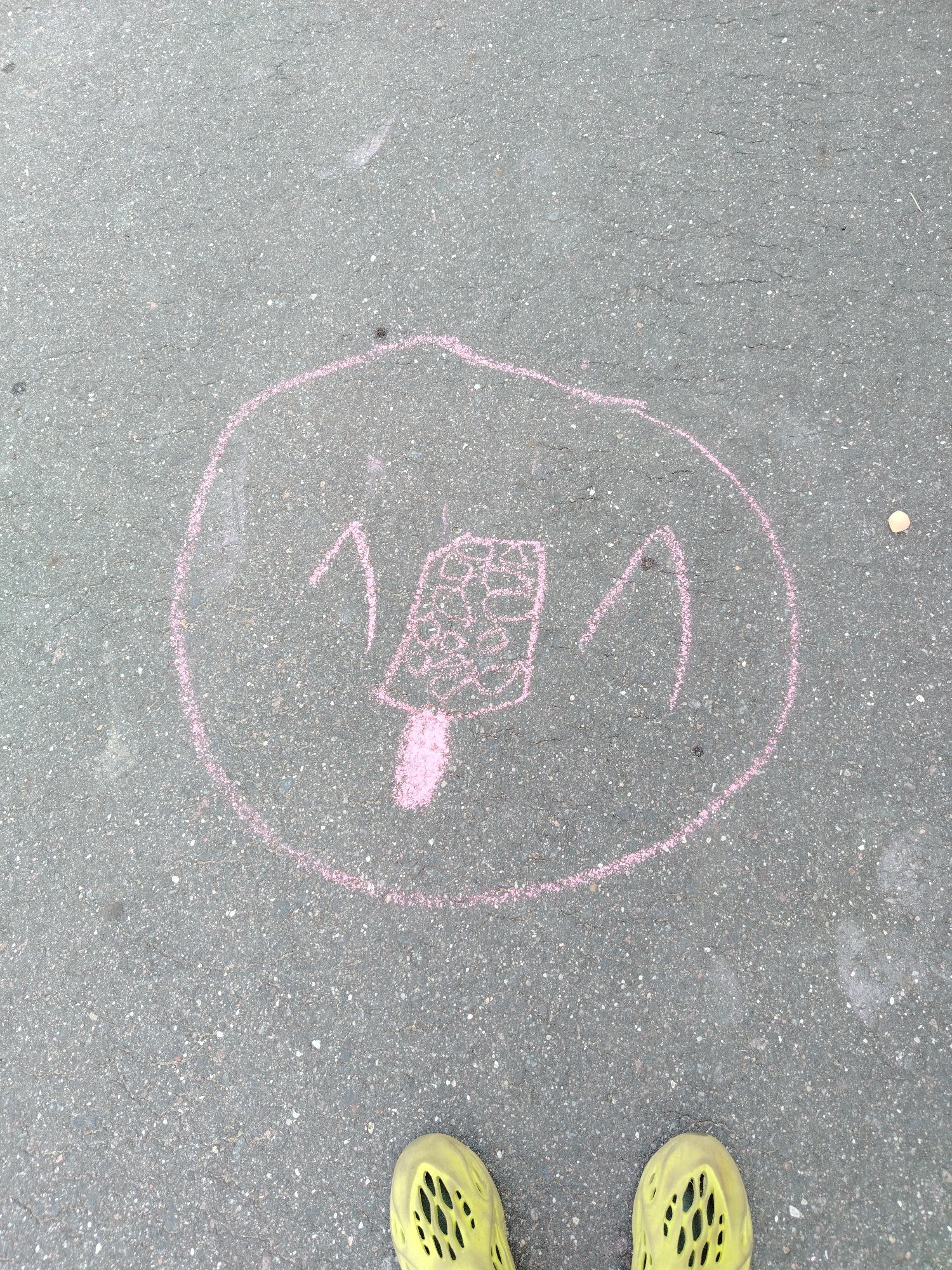 The image shows a chalk drawing on asphalt. The drawing is contained within a pink circle and depicts a stylized popsicle with a face. Two upward-pointing V shapes suggest ears or eyes. The popsicle itself is drawn with a grid pattern. Two yellow shoes are visible at the bottom of the frame, suggesting a person is standing over the drawing. The environment appears to be an outdoor urban setting, likely a sidewalk or street.