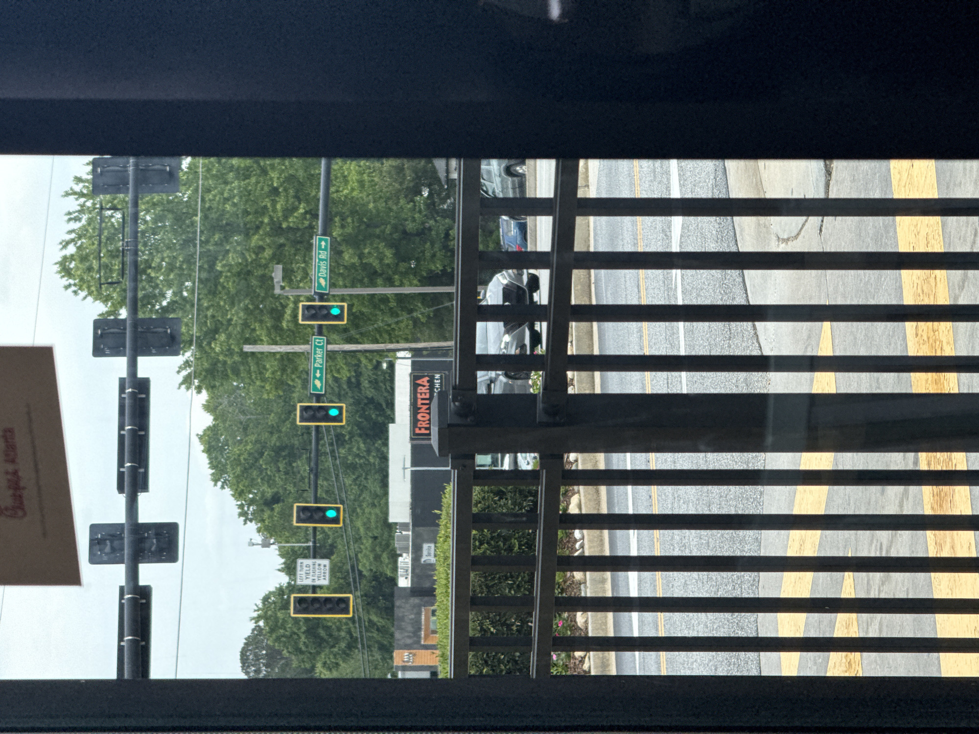 The image shows an exterior view through a window in Stone Mountain, Georgia, USA. A pole supports street signs that read "Parker Ct" and "Davis Rd". Traffic lights are on the pole with a green light illuminated. A sign reads "LEFT TURN, YIELD ON FLASHING YELLOW ARROW" and a business sign reads "FRONTERA KITCHEN". A road and yellow median can be seen, and the bars of a railing are visible. Cars are parked along the street.