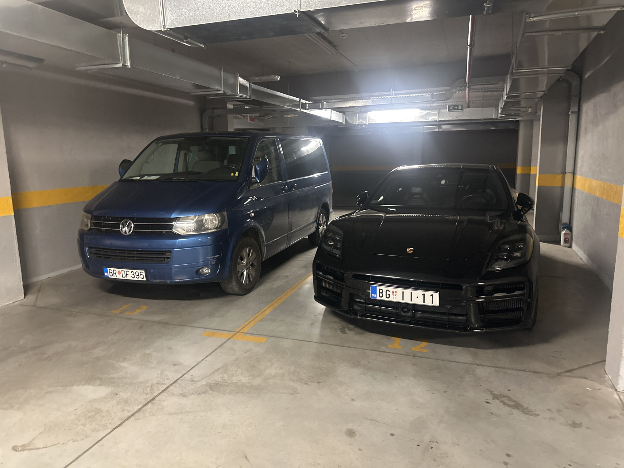 This media file captures a dimly lit indoor parking garage, likely in Boreti, Montenegro. The scene features two cars parked side-by-side in designated spots marked with yellow lines and numbers. On the left is a blue Volkswagen minivan. Its license plate is visible and reads "BR DF 395". To the right of the minivan is a sleek black Porsche. Its license plate displays "BG 11-11". The lighting suggests it is either nighttime or the garage is not well-lit. The environment appears to be a typical concrete parking structure with exposed ventilation systems running along the ceiling. No people are visible in the frame. The overall impression is one of orderly storage of vehicles within a functional, utilitarian space.