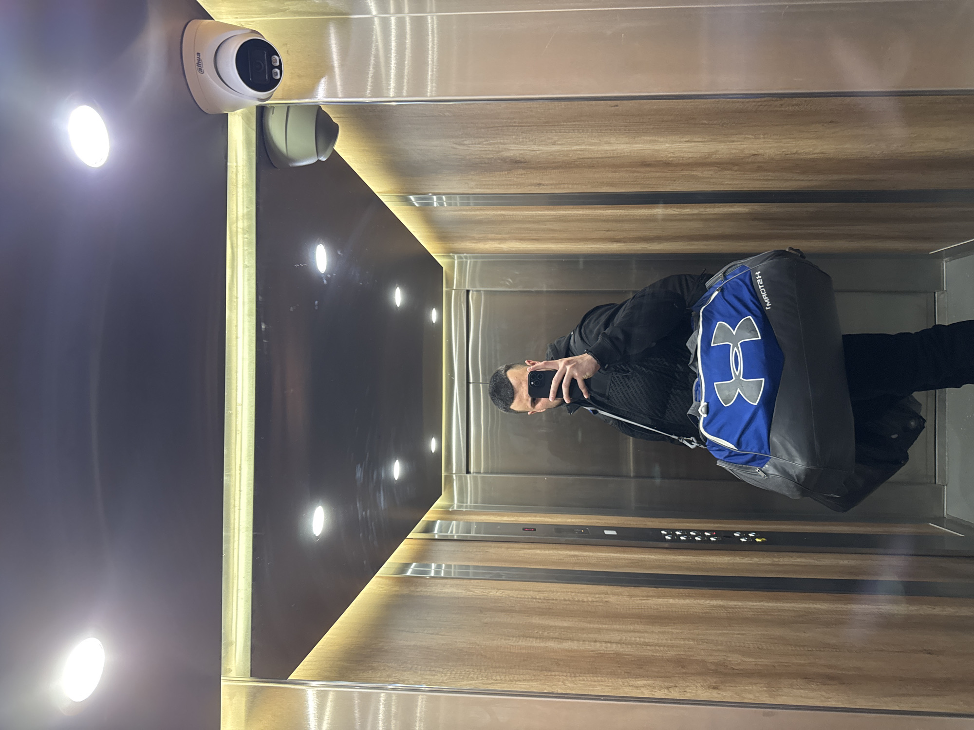 A man is taking a selfie in an elevator with light brown wooden and stainless steel walls. The elevator has a security camera in the corner, as well as a row of lighted buttons. The man is wearing a black jacket and carrying a blue and gray Under Armour bag. The location appears to be an elevator, but further information to establish the location in the city of Sofia, Bulgaria, is not available from the image.