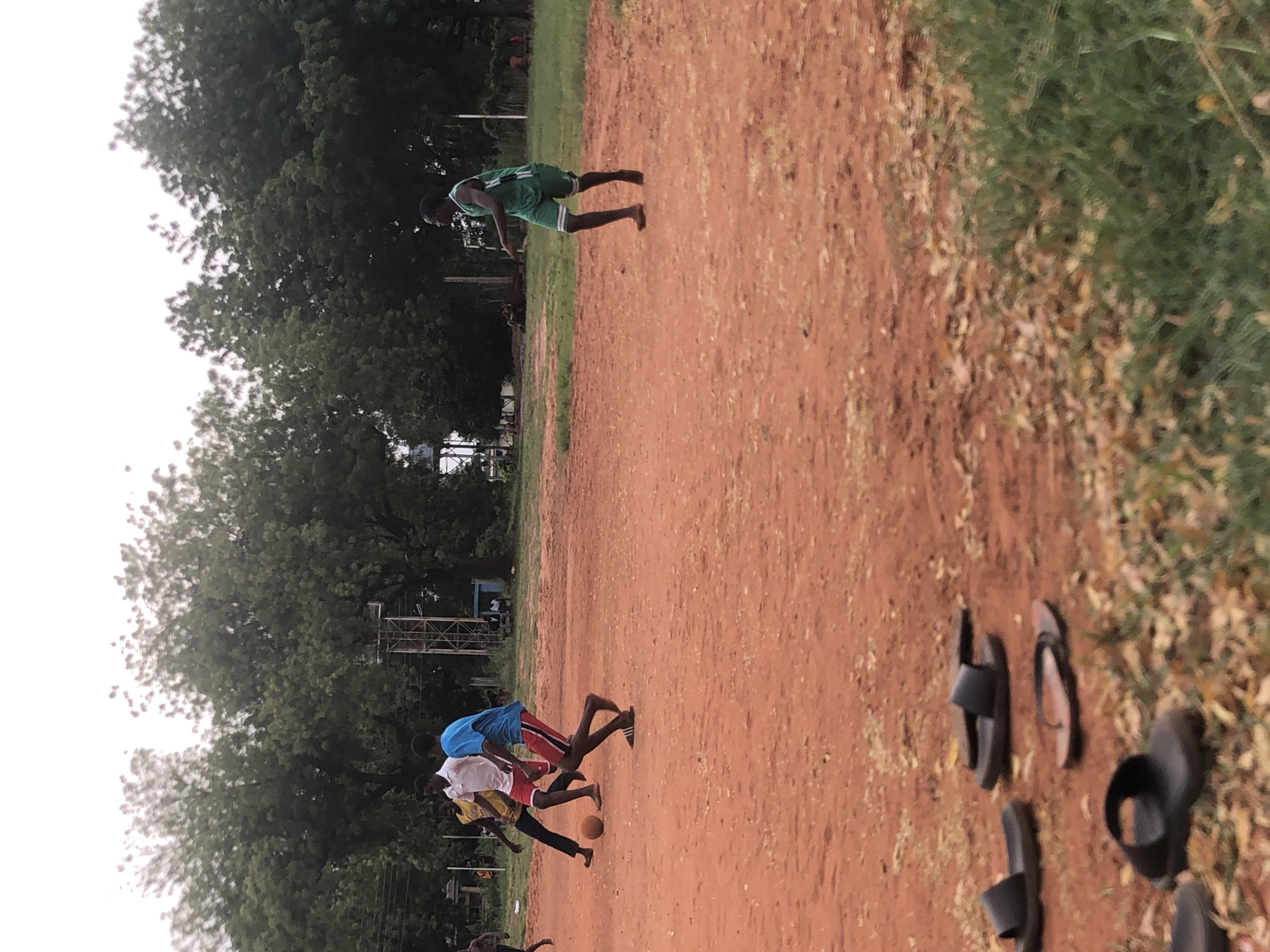 This image, captured in Ukwu-Abwa, Nigeria, depicts an outdoor scene featuring several young males engaged in recreational activity on a reddish-brown dirt field.
In the mid-ground to the left, a young man wearing a green sleeveless top and matching shorts is shown in a dynamic, leaning-forward posture, suggesting he is running or about to move. Closer to the center of the frame, three other young males are actively involved in a game of football (soccer). One is identifiable by a blue t-shirt and red shorts, another by a white t-shirt and red shorts, and a third is partially visible in a yellow t-shirt and dark pants. A brown football lies on the ground near this group, indicating ongoing play. Other blurry figures are faintly visible in the background and foreground.
The ground is predominantly reddish-brown earth, with scattered patches of green grass and dried leaves, especially along the edges. The background is filled with a dense canopy of tall, green trees, under a bright, softly lit sky, possibly indicating an overcast day or late afternoon. Some faint structural elements, resembling scaffolding or building frames, can be seen through the trees in the distance. In the foreground to the far right, several pairs of dark-colored flip-flops or sandals are scattered on the ground, suggesting players have removed their footwear. The scene conveys a casual and active environment. No visible text is present in the image.