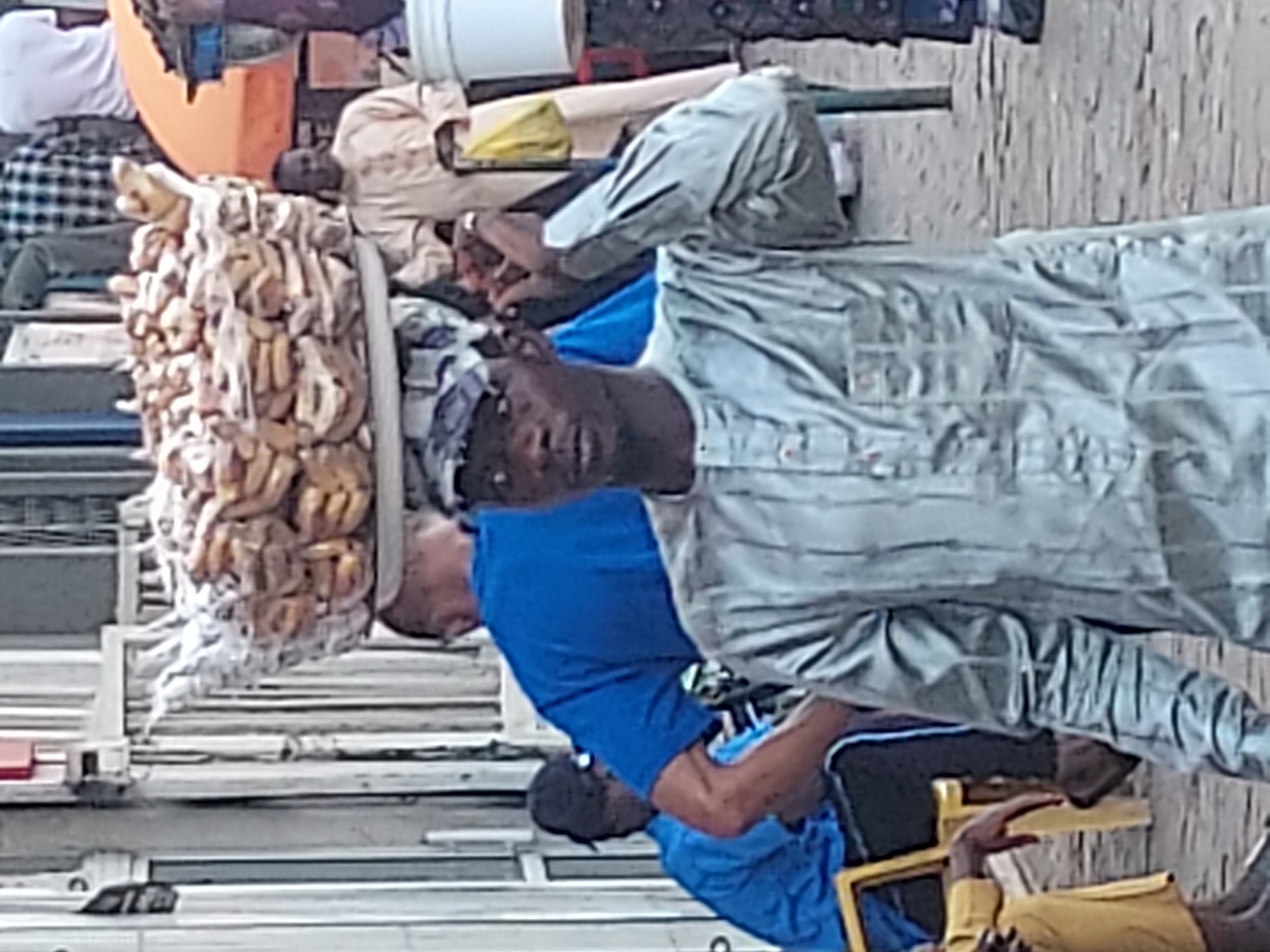 In Jos, Nigeria, a vendor stands near a display of bundled smoked fish. Three men are visible: one wearing a blue shirt, one a gray striped shirt, and another wearing a yellow shirt. A white bucket and other unidentified objects are in the background. The scene appears to be outdoors, possibly near a street or market. The setting is daytime.