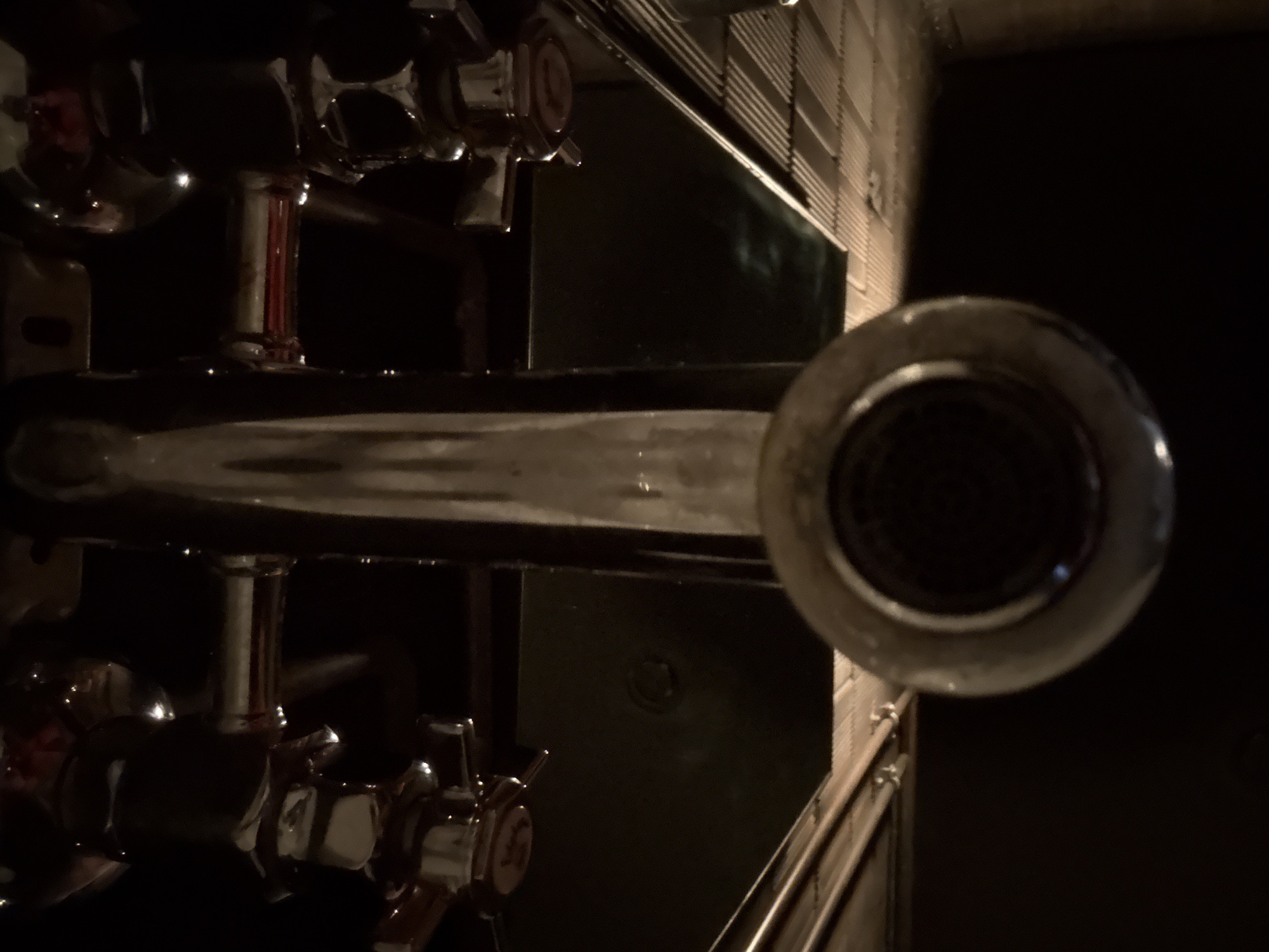 The image depicts a dimly lit indoor scene, likely a bar or pub, viewed from a slightly high angle and rotated 90 degrees clockwise.
In the prominent left portion of the image, multiple chrome-plated dispensing taps are visible, suggesting a drink-serving setup. These taps are highly reflective, catching faint highlights from the ambient light. On the lever of one tap, a subtle red marking, possibly a stylized letter or logo, can be discerned.
Below the taps, a dark, flat, and somewhat reflective surface, resembling a bar counter, extends horizontally across the frame. On this surface, in the center-right area, there is a faint circular indentation or detail.
On the far right, a large, dark, circular object, possibly a lens or a cap of some machinery, partially fills the lower right corner, its surface showing some texture or grid pattern in its center.
The background on the right side features a textured wall with distinct horizontal lines, which could be patterned tiles or paneling, receding into the darkness. The overall lighting is very low, creating a subdued atmosphere, characteristic of an evening or nighttime setting. No individuals are present in the scene. The visual cues strongly indicate a commercial establishment like a bar.