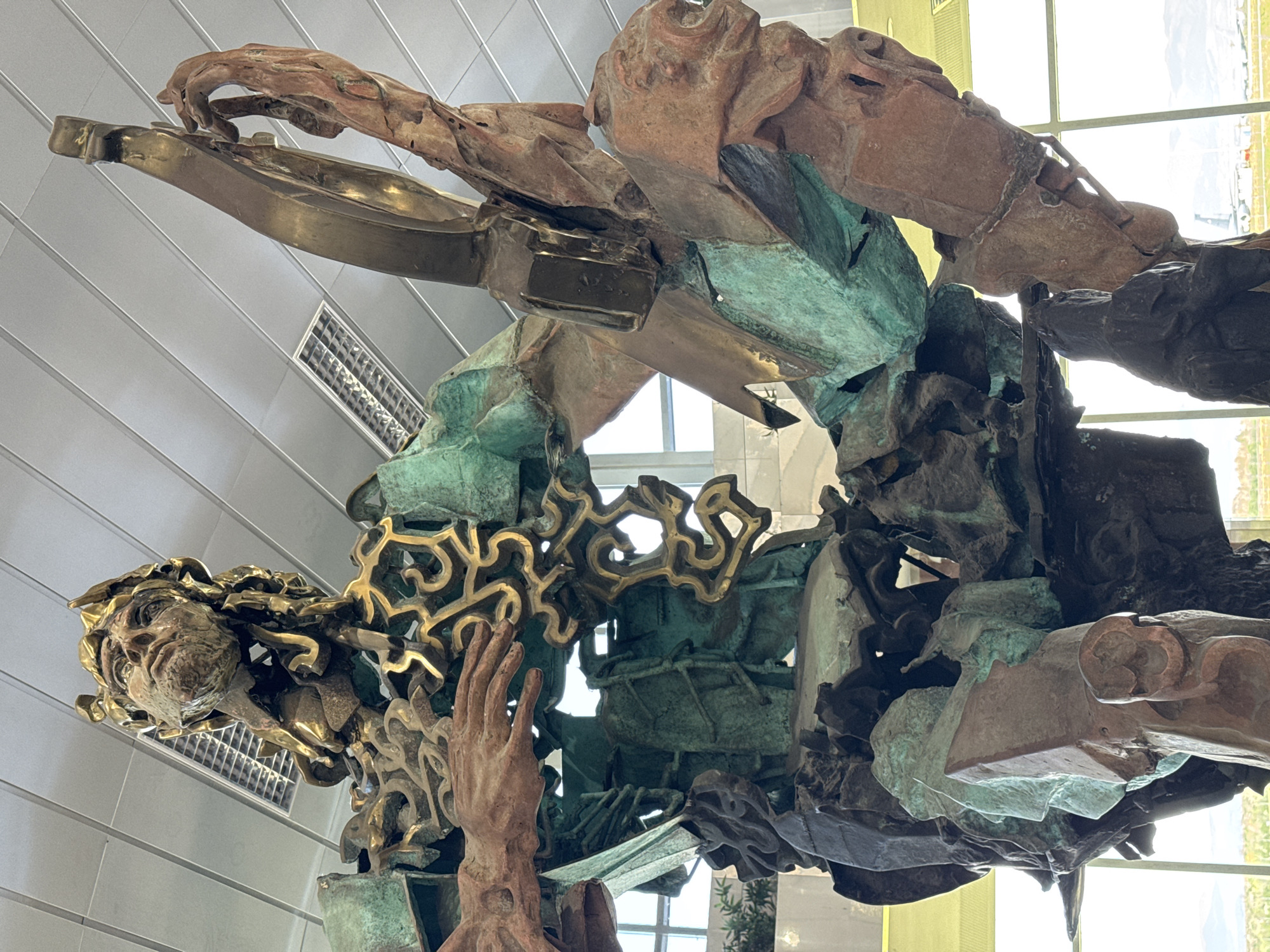 The image displays a large, complex sculpture from a low, upward-looking perspective, rotated counter-clockwise. The artwork is a blend of materials, primarily featuring golden bronze, reddish-brown metallic components, and significant sections with a distinctive verdigris green patina, indicative of oxidized copper or bronze.
The sculpture portrays fragmented human-like forms intertwined with abstract and symbolic elements. A prominent stylized human face, with intricate golden-colored hair or flowing patterns, is visible on the lower left, conveying a sense of intensity or thought. Nearby, a reddish-brown hand reaches upwards. Another reddish-brown hand is depicted interacting with a golden bronze structure resembling a keyboard or piano, hinting at themes of music, creation, or communication.
The composition is dynamic and dense, combining angular geometric forms with organic, swirling golden patterns. The fragmented limbs and body parts appear to be either emerging from or dissolving into the intricate metallic mass. The sculpture evokes a sense of motion, struggle, or transformation.
The artwork is situated indoors within a modern architectural setting. A light grey, paneled ceiling with rectangular vent or light fixtures is visible. Large windows in the background allow abundant natural daylight to illuminate the sculpture, suggesting it is daytime. The bright light also reveals a blurred outdoor environment beyond the windows. No discernible text is visible on the sculpture or in the background. The scene suggests a public space in Sofia, Bulgaria.