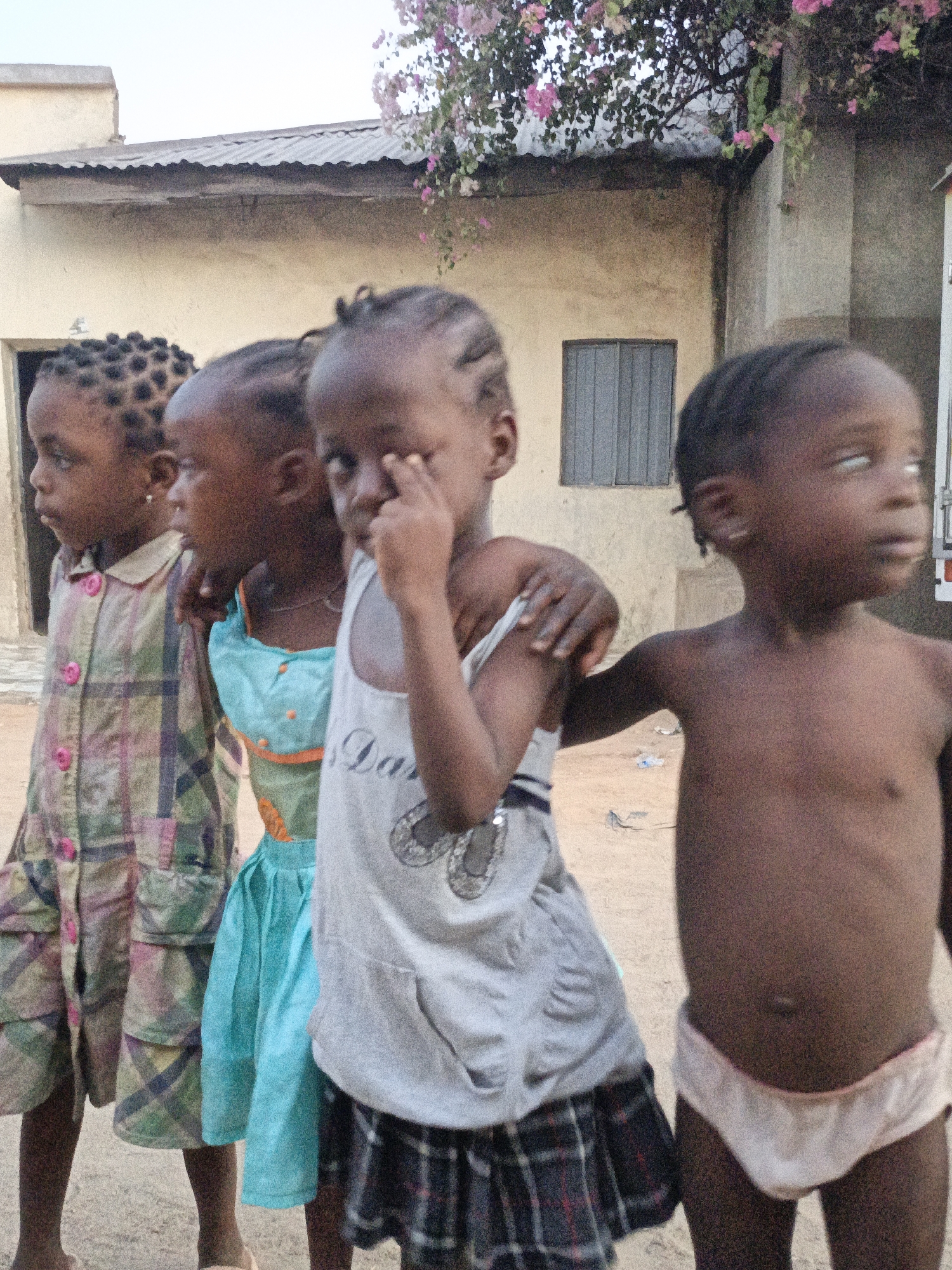 Four young children stand shoulder to shoulder in an outdoor setting in Gwagwarwa, Nigeria. The first child, seen from the left, wears a multi-colored plaid shirt and shorts. The second child has a light blue dress. The third child is in a white tank top and a plaid skirt, with her fingers near her eye. The fourth child is shirtless except for light-colored underpants. Behind them is a building with a corrugated metal roof and a single window, and a flowering vine drapes above. The ground appears to be dirt.