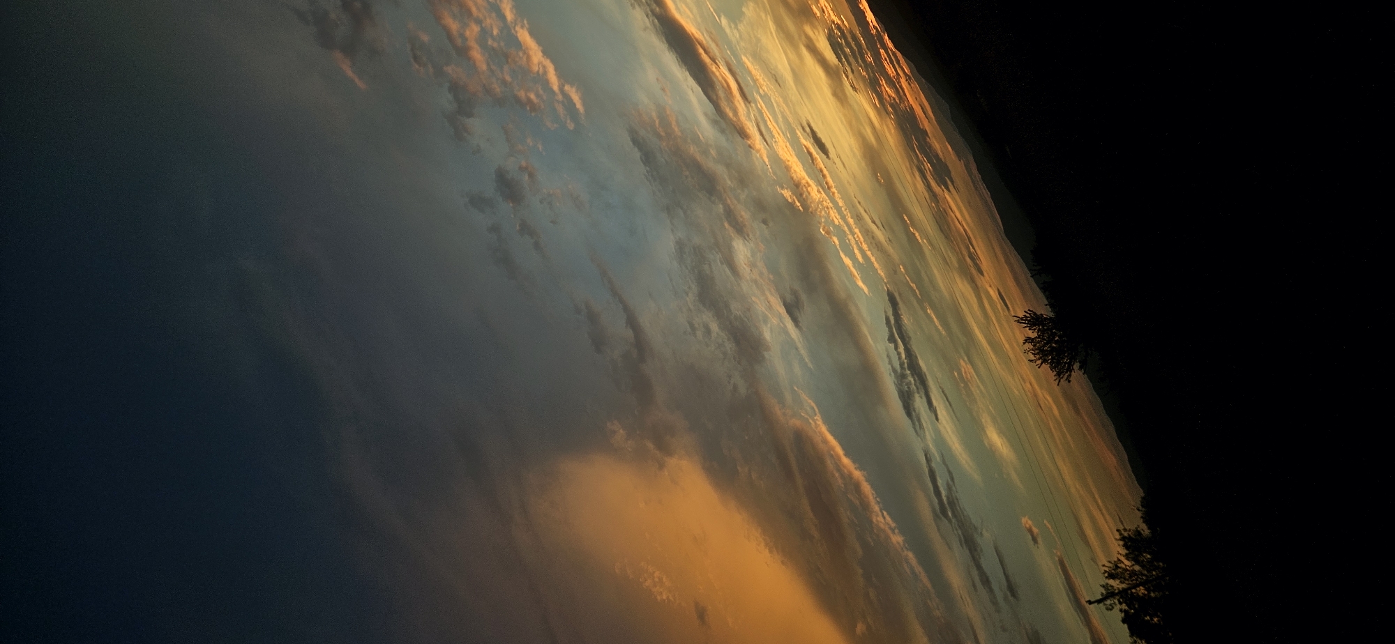 A photograph captures a dramatic sunset over a silhouetted landscape. The sky is a gradient of deep blue at the top, transitioning through shades of gray, teal, and finally a vibrant orange and yellow near the horizon. Wispy clouds are scattered across the sky, illuminated by the setting sun, creating streaks of light and shadow. In the foreground, the land is entirely black, obscuring any details of terrain or vegetation, except for a few dark shapes that might be trees. The overall impression is one of a vast and awe-inspiring natural spectacle, with the earth appearing as a dark, indistinct mass against the vibrant sky. The location is Momin Prohod, Bulgaria.