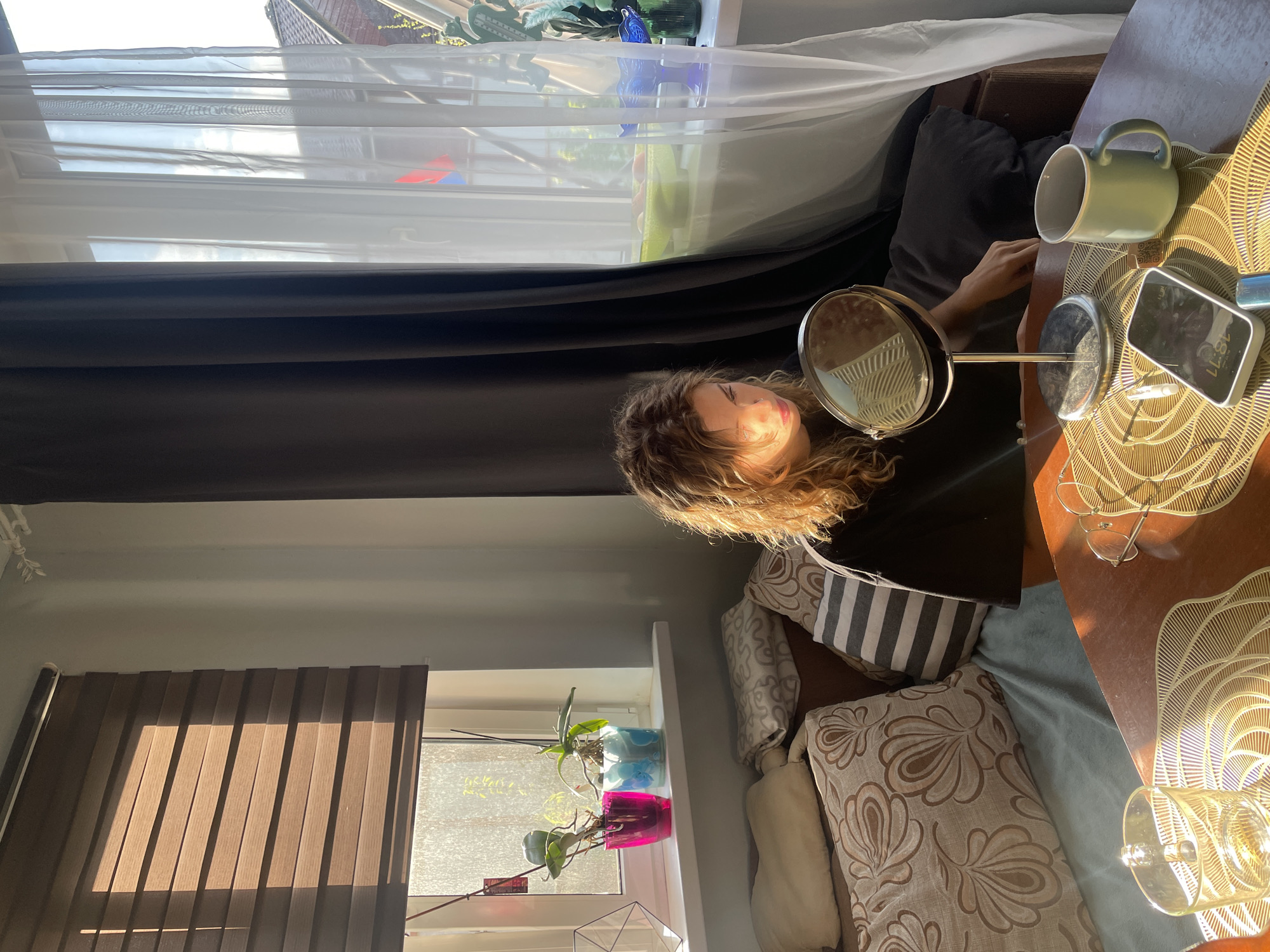 The image appears to be taken indoors in a room with two windows. One window has vertical blinds and the other is draped with a light-colored sheer curtain overlaying a dark curtain. A person is seated, looking into a round magnifying mirror on a stand. A light green mug, a cell phone displaying the time "18:11", and a pair of eyeglasses rest on a brown surface covered by a gold-colored decorative placemat. Several pillows are behind the person. Two plants in colorful pots are on the window sill by the blinds. The scene takes place in Sestroretsk, Russia.