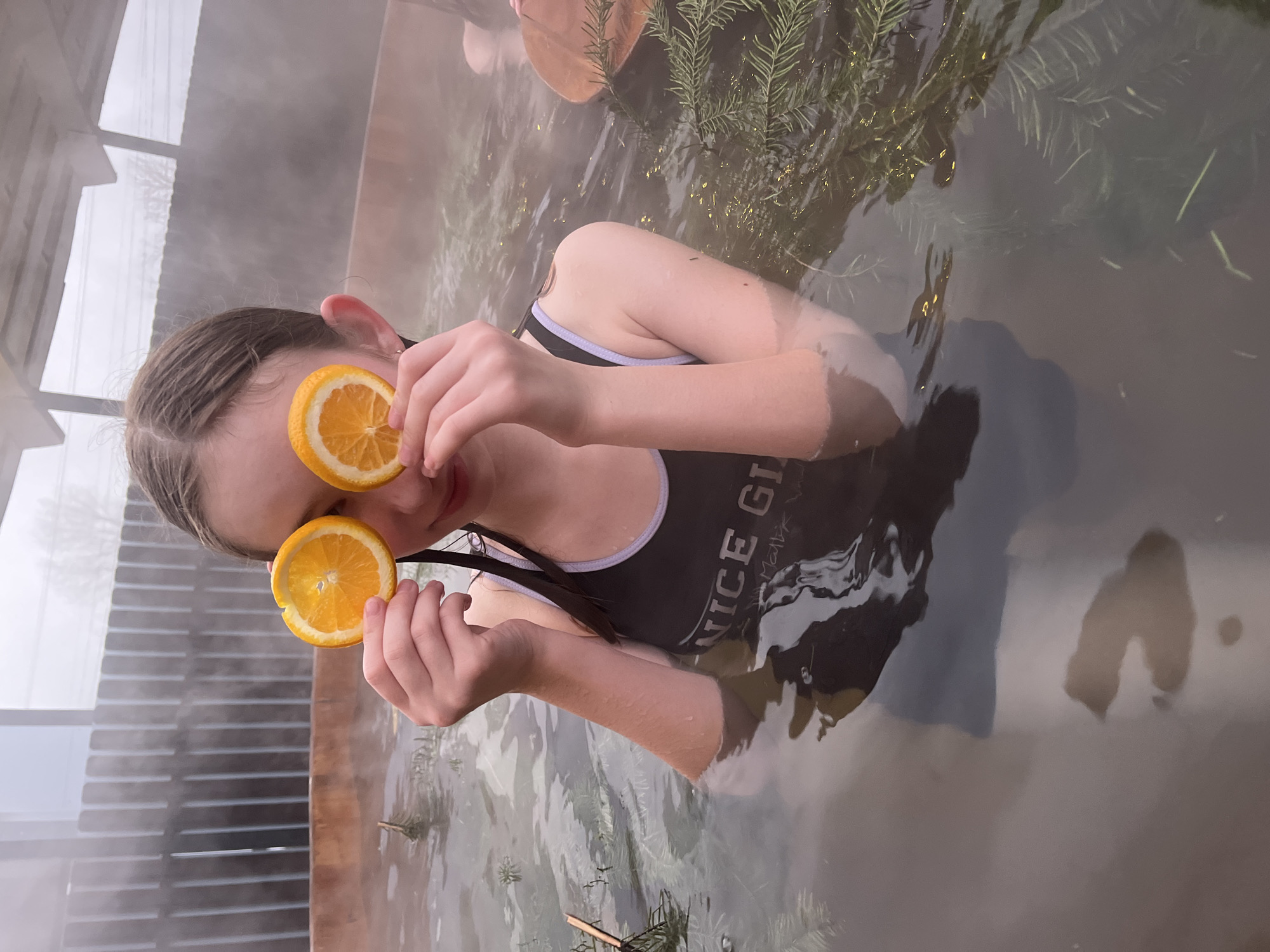 A young girl is in a body of water, holding orange slices over her eyes. She is wearing a black swimsuit with a light-colored trim, and her hair is pulled back. There are pine boughs floating in the water. The scene appears to be a hot tub or similar bathing facility, possibly located outdoors. There is steam rising from the water. In the background, there's a wooden structure that could be part of a building or enclosure. The location in the city is unknown.