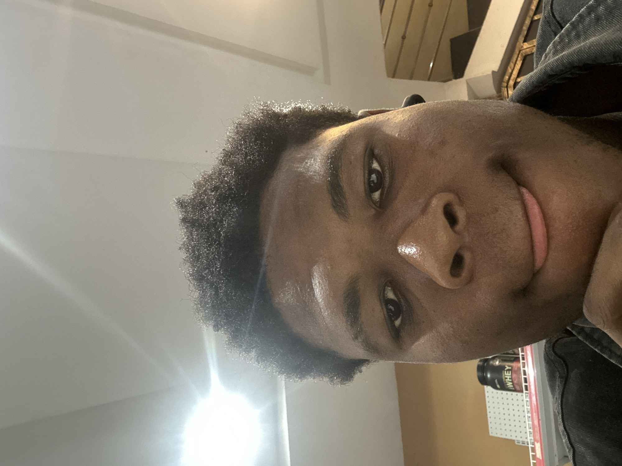 A close-up, low-angle selfie shows a young Black person's face. Their eyes are open, looking towards the camera, and their mouth is slightly open with the tongue sticking out. The person is wearing a dark denim jacket, and one hand is visible resting near their chin. Behind them, a shelf holds a black and red container labeled "WHEY." The background also shows a staircase and the ceiling, which is illuminated by bright lights. The image appears to have been taken indoors in a public or commercial space.