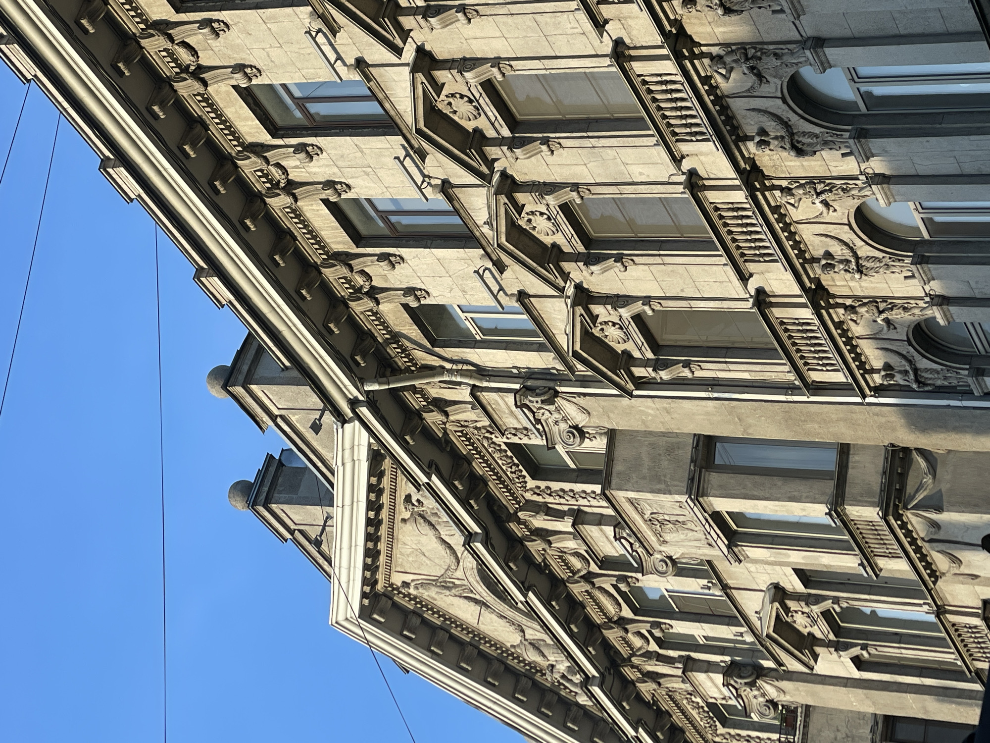 The image displays a low-angle, upward view of an ornate building facade against a clear, deep blue sky, characteristic of architectural styles found in Saint Petersburg, Russia. The perspective is rotated counter-clockwise, presenting the building's intricate details diagonally across the frame.
The building's facade is constructed from light-colored stone or stucco, likely in shades of grey and beige, and features multiple stories with large, recessed rectangular windows. Each window is surrounded by elaborate architectural ornamentation, including volutes, rosettes, and shell motifs, particularly visible in the cropped images. Projecting cornices and ledges separate the floors, supported by numerous decorative brackets and corbels. Many of these supports are sculpted with grotesque-like faces, stylized animal heads (resembling lions or mythical creatures), and other classical elements, creating strong three-dimensional relief. Further details include intricate relief carvings integrated into the facade panels, some depicting figures or abstract patterns, and decorative false balustrades beneath windows.
Strong, bright sunlight illuminates the facade, casting pronounced shadows that highlight the depth and complexity of the carvings and architectural elements. The sky is entirely cloudless. Thin, dark utility wires or cables traverse the upper left portion of the image against the blue sky. There is no discernible activity taking place, and no people or visible text are present in the frame. The overall impression is one of grand, historical architecture captured in sharp detail under bright daylight.