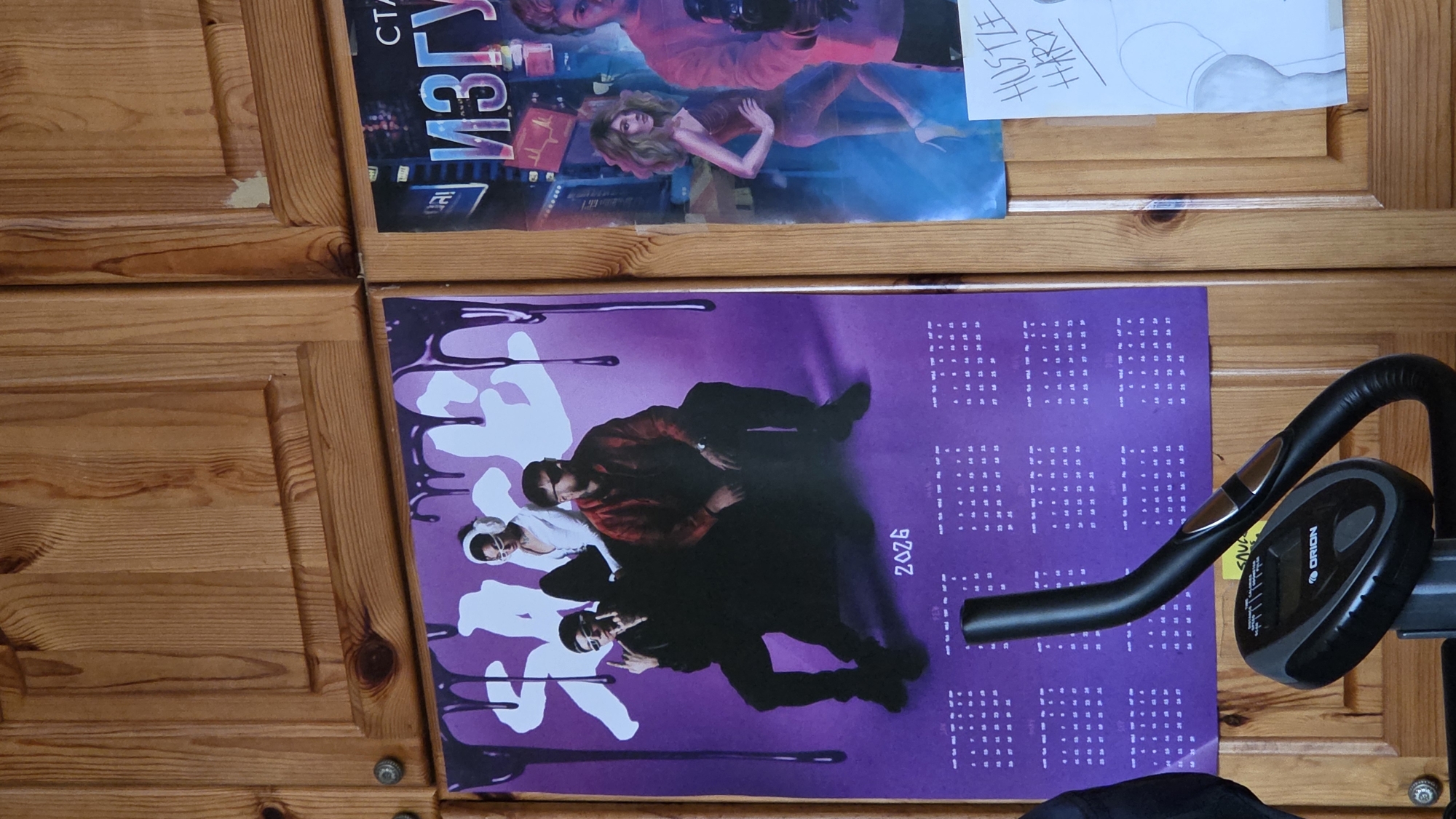 The image shows a close-up, eye-level, indoor shot of a wooden cabinet. Hanging on the cabinet doors are two posters and a drawing. The poster on the left features three people dressed in casual clothing. The person in the middle is wearing headphones and a white top, while the two people on either side are seated and wearing dark clothing. The poster has a purple background with a dripping effect at the top and the year "2026" printed near the bottom. The poster on the right is partially visible and features an image of a woman in a red dress. To the right of this poster is a drawing of a person's legs and buttocks. In the foreground, a black exercise bike is positioned such that its handlebars and console are visible. The environment appears to be a room with wooden paneling.