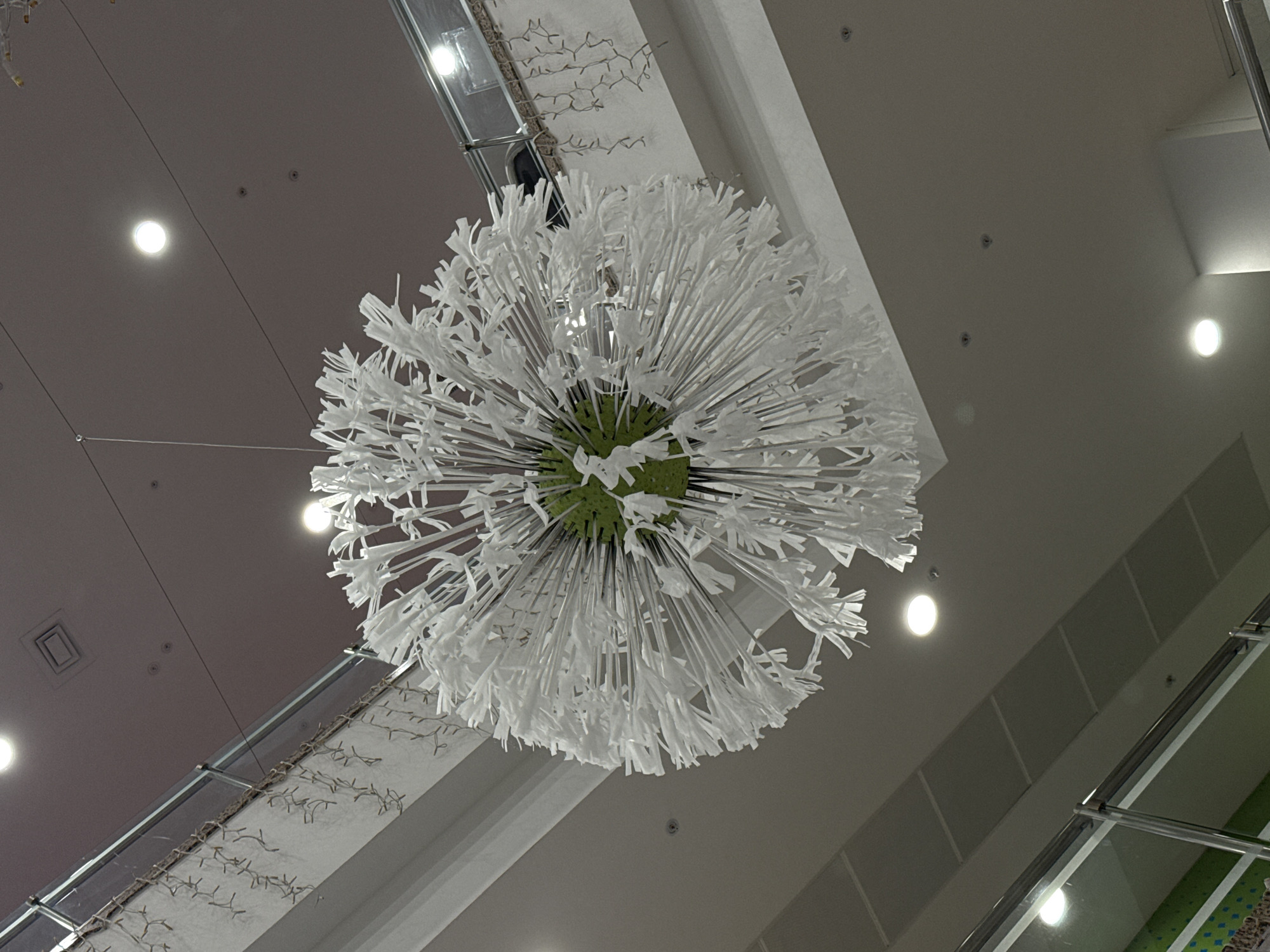 The image displays a large, spherical decorative fixture suspended from a high, light gray ceiling in what appears to be a modern indoor space. The fixture itself resembles an oversized white dandelion or an allium flower, characterized by a central green core from which numerous thin, white, frond-like elements extend outwards in a radial pattern, creating a fluffy and voluminous form. These white elements appear to be made of a lightweight material, possibly paper or fabric, cut into varied, ribbon-like shapes.
The ceiling is adorned with multiple recessed circular spotlights, casting bright, diffused light. Other smaller dark circles are also visible on the ceiling, likely additional fixtures or structural elements. In the upper right and lower right portions of the frame, metallic structural beams or frames are visible. Adjacent to these, a wall or architectural surface features an intricate pattern of thin, branched elements, possibly part of a decorative lighting arrangement or an artistic installation, given the presence of small, bright points within the pattern, suggesting tiny lights. Reflections on smooth surfaces hint at glass partitions or railings within the expansive interior.
No people or activities are visible, and no text is present in the image. The setting suggests a public or commercial building, such as a shopping mall or an atrium, illuminated entirely by artificial light.