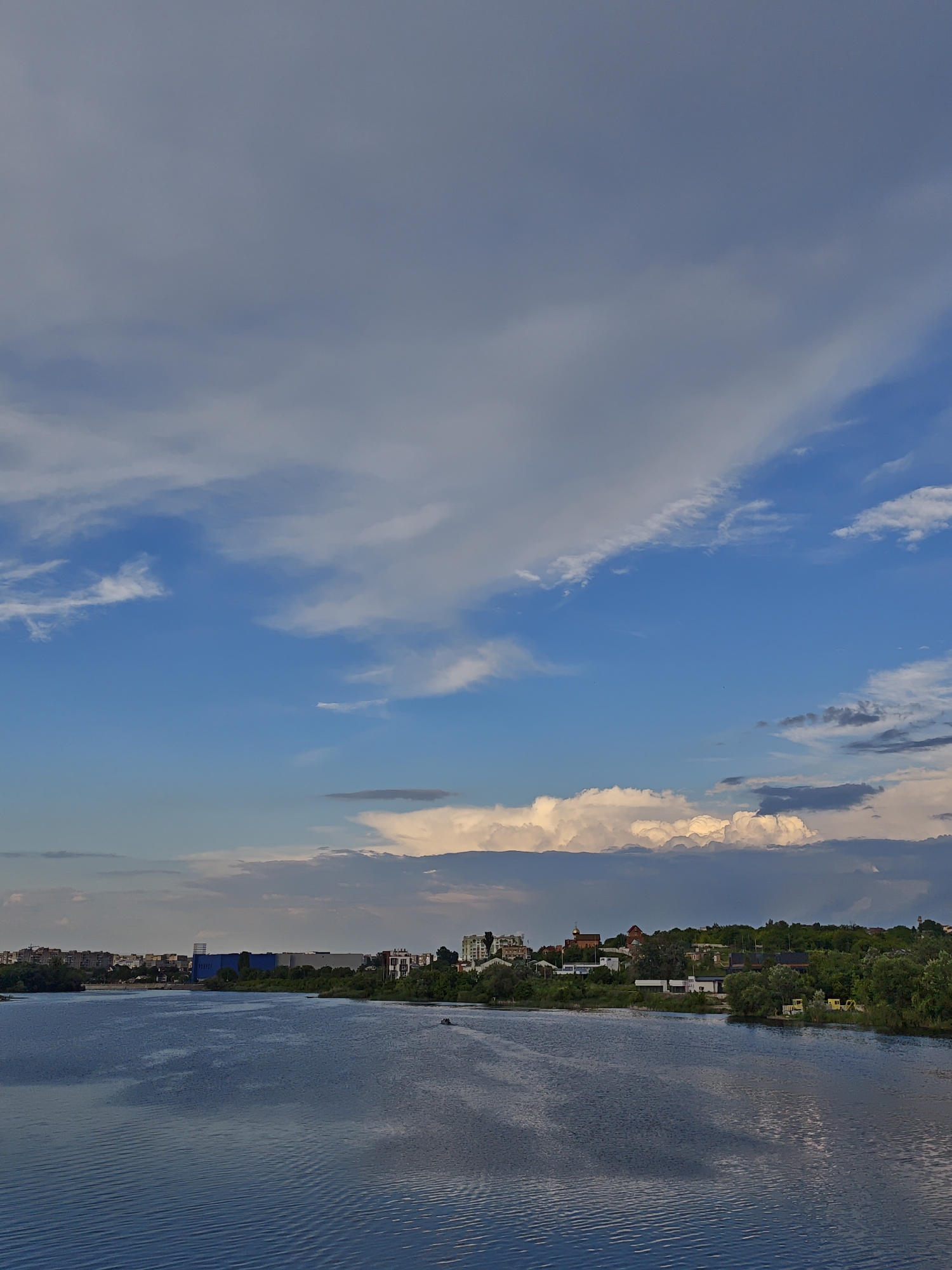 The image depicts a wide, calm river flowing through an urban landscape under a partly cloudy sky. The water reflects the sky, creating a muted blue and grey surface. Along the riverbank, there is a dense line of green trees, interspersed with buildings of various sizes and architectural styles, suggesting a populated area. Some of these buildings appear to be residential, while others are larger, possibly commercial or industrial structures. In the distance, the silhouette of a city is visible against the horizon. The sky is a dynamic mix of bright blue patches and large, dramatic clouds, some of which are illuminated by sunlight, giving them a warm, golden glow. The overall impression is one of a serene, yet active, waterfront setting.
This scene is likely the Southern Bug River in Vinnytsia, Ukraine, as it flows through or near the city, with the buildings on the far bank representing the urban development along the river.