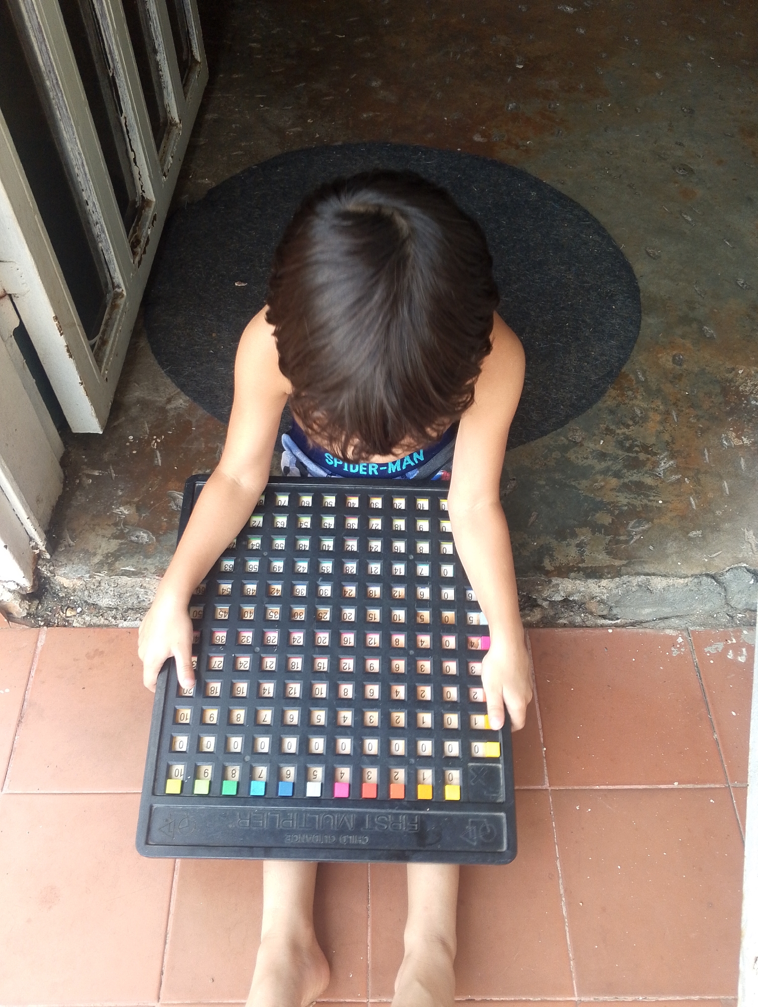 A young child sits on a brown tiled floor, holding a "First Multiplier" educational toy. The child is wearing a blue Spider-Man tank top. The toy is a black board with numbered tiles, set up for multiplication practice. Behind the child is a gray doormat and a white, weathered door frame. The child is barefoot. The location of this image is not known to be San Carlos, Venezuela.