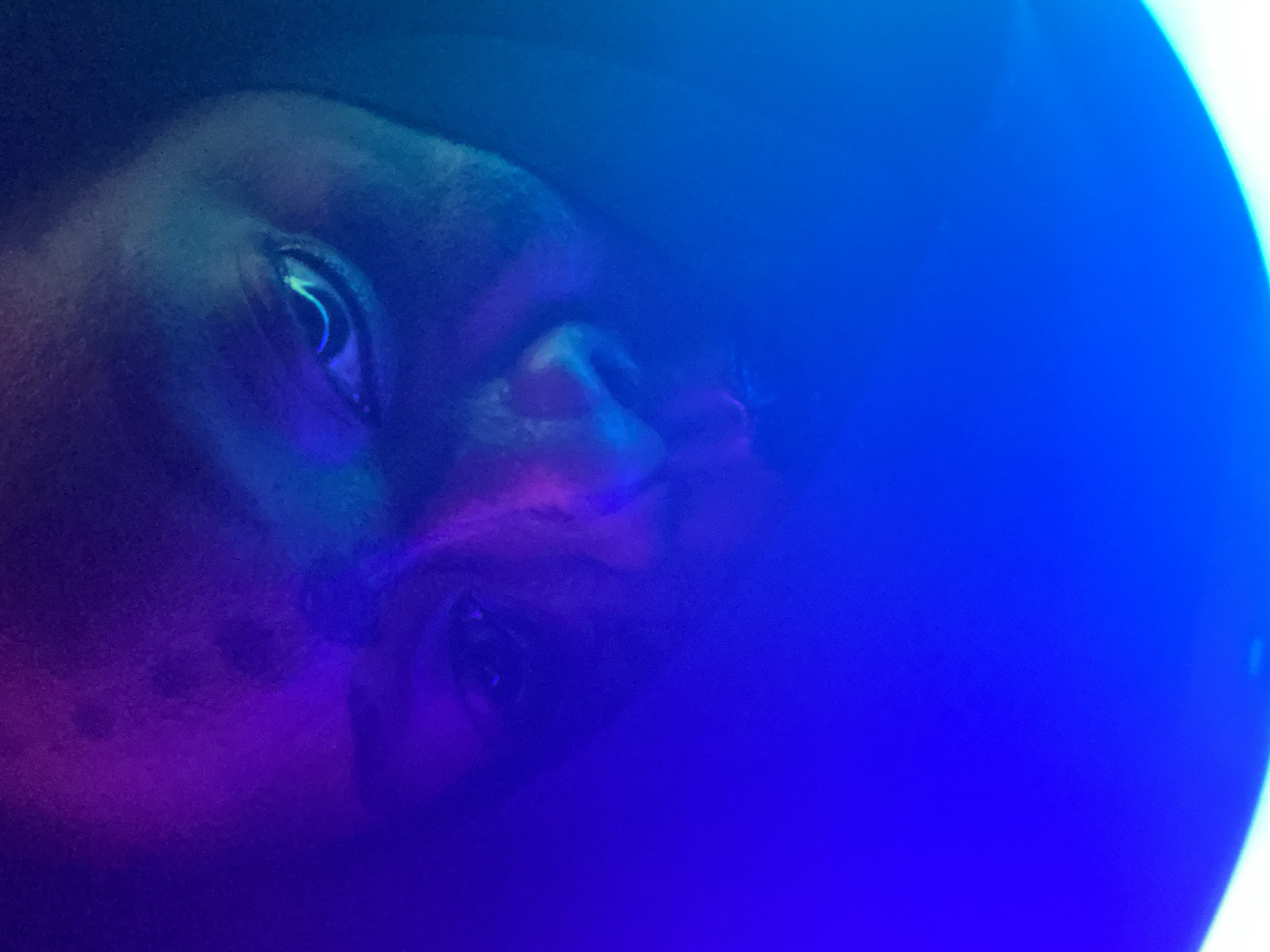 A close-up shot reveals a face, likely male, bathed in predominantly blue and purple hues. The facial features, including the eyes, nose, and mouth, are visible, though partially obscured by the colored lighting. The face is turned slightly to the side, with the top of the head cropped out of the frame. The environment is undefined, with the focus solely on the face and the lighting effects. The image does not provide any information that allows to determine the location in the city of Maigizo, Nigeria.