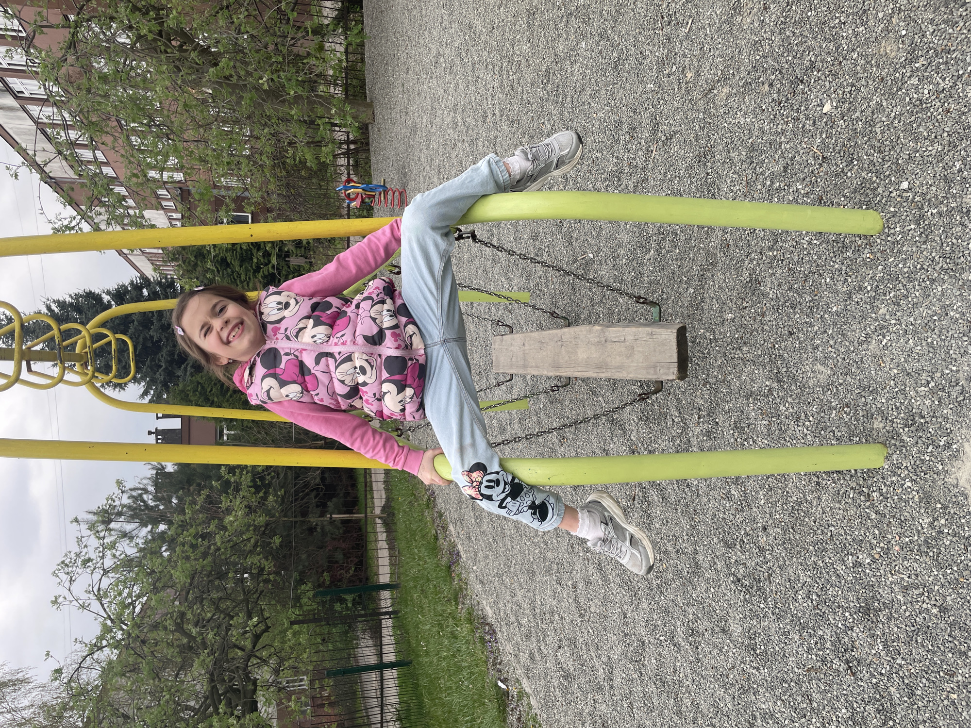 A girl in a pink vest and blue jeans is sitting on a yellow bar in a playground. The ground is covered in gravel. There are trees and a building in the background.