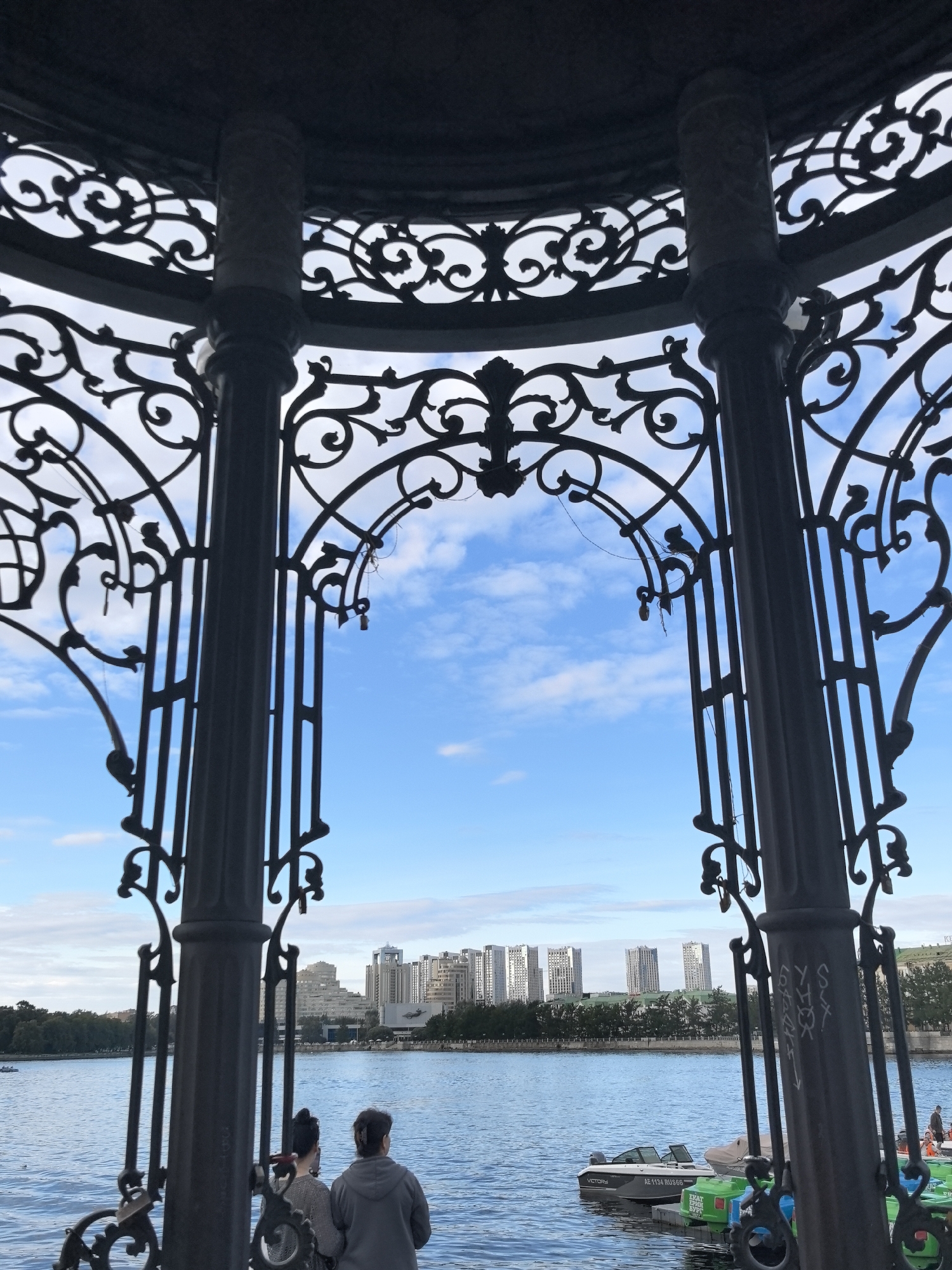 The image, taken from within a decorative metal gazebo, looks out over a body of water towards a city skyline. The gazebo features intricate black metalwork with spiral motifs and decorative columns. In the foreground, two individuals, viewed from behind, are seated facing the water. The water is a vibrant blue, with small ripples indicating a gentle breeze. Several small boats, some with green hulls, are docked along the water's edge. Across the water, a row of modern buildings, characterized by their white facades and uniform window patterns, dominates the horizon. Trees line the waterfront between the water and the buildings. The sky is a clear blue with scattered white clouds. The scene is likely in Yekaterinburg, Russia, given the architecture of the buildings and the general appearance of the waterfront.