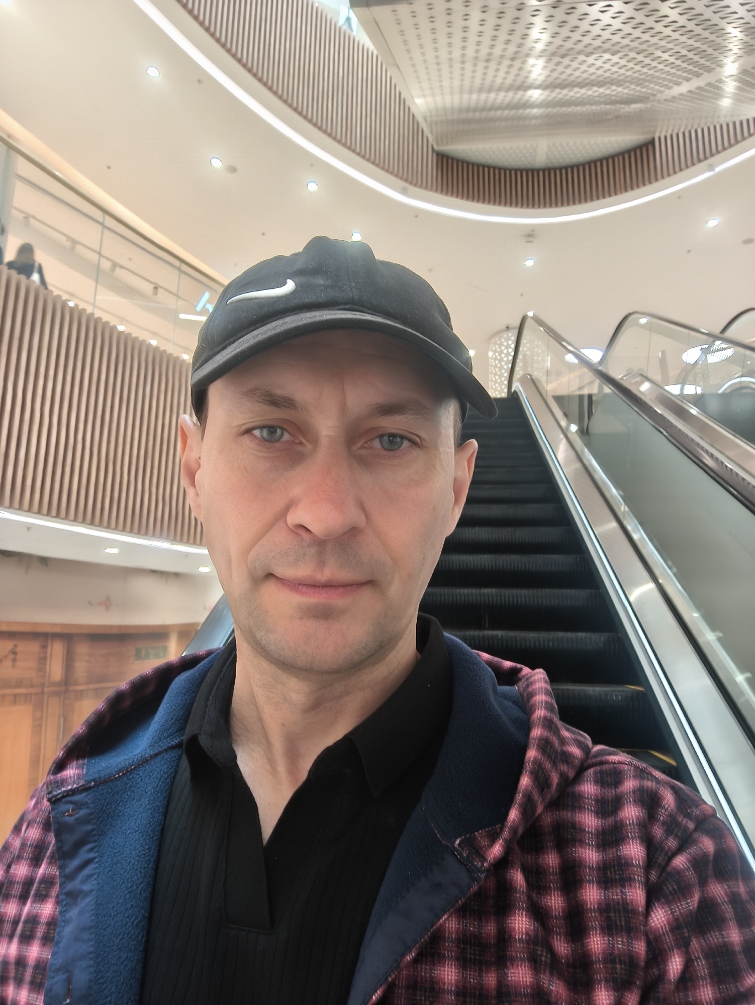 A man wearing a black baseball cap with a white Nike logo, a black collared shirt, and a red and black plaid jacket is taking a selfie in a modern indoor space, likely a shopping mall or airport, in Vladivostok, Russia. He has blue eyes and short, light brown hair. Behind him, an escalator is visible, with sleek glass railings and dark treads. The background features architectural elements such as vertical wooden paneling and a multi-tiered ceiling with integrated lighting. The lighting is bright and diffused, suggesting an interior environment. The man is positioned centrally in the frame, with his face dominating the upper portion.