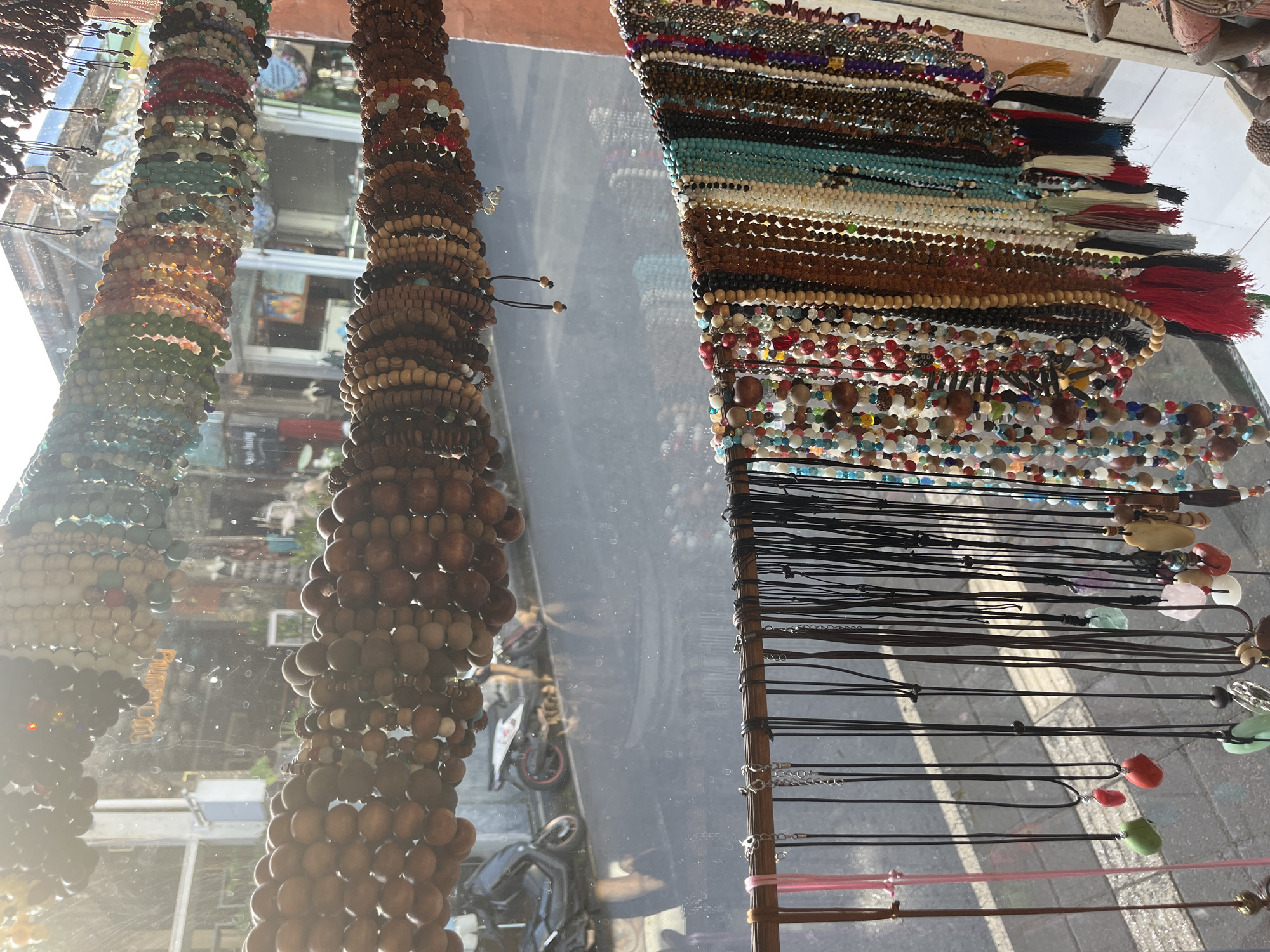 The image shows a display of jewelry, seemingly viewed through a storefront window, in what appears to be the Ubud District, Indonesia, given the styles of the merchandise and the architectural elements visible in the reflected street scene.
On the left, multiple strands of beaded bracelets, made of colored stones and wood beads, hang vertically. In the center, a similar display features a collection of brown wood beaded bracelets. On the right, necklaces hang vertically from a rack. These necklaces consist of beaded strands with tassels and gemstone pendants on cords.
The street outside is visible as a reflection in the glass. Motor scooters are parked on the street, and architectural details of buildings across the street, including signage, are discernible. The light suggests it is daytime.