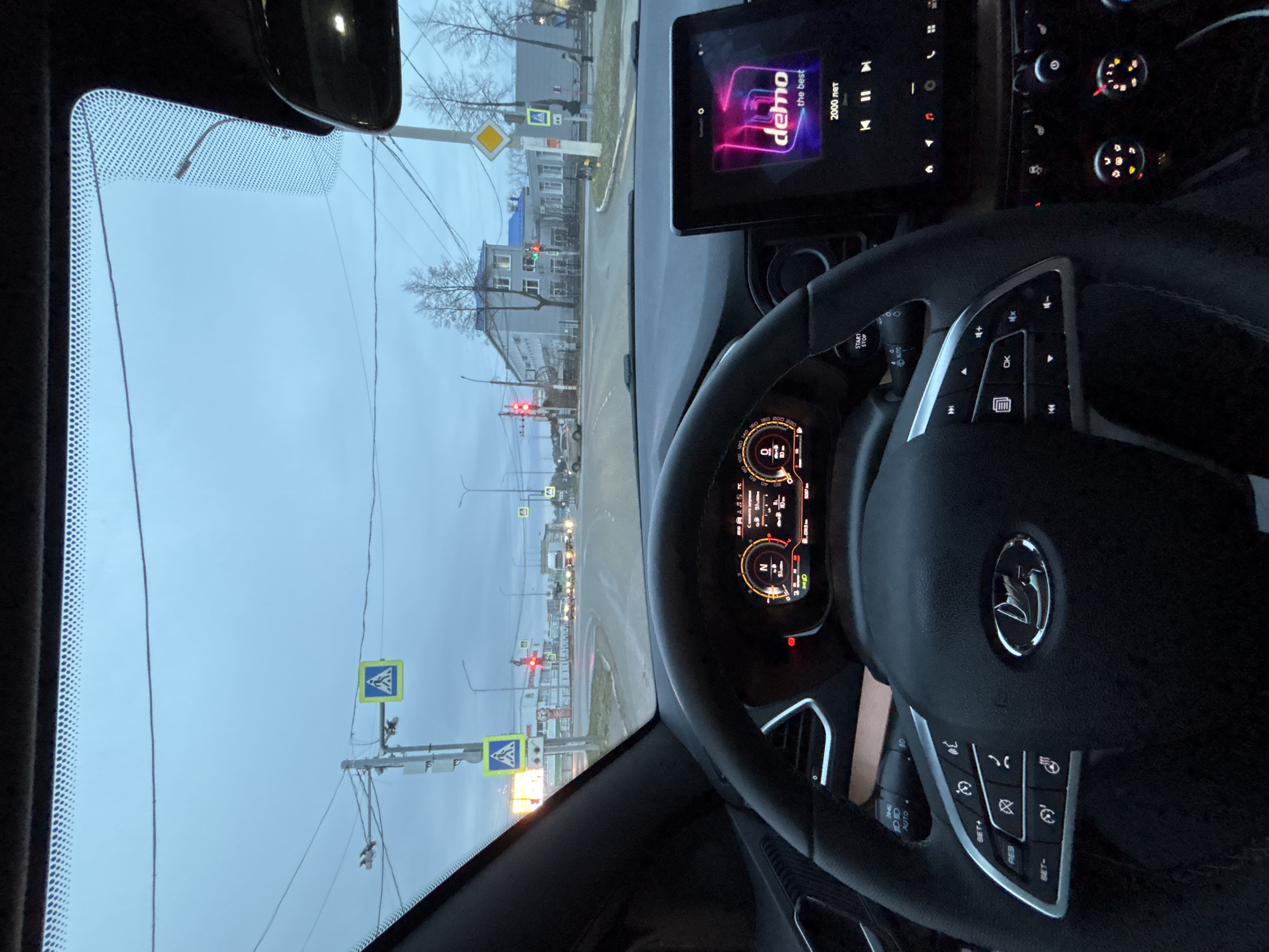 Here's a forensic description of the image:
**Scene:** Interior of a vehicle, likely a car, looking through the windshield. The location appears to be an urban area, possibly Chaikovsky, Russia.
**Environment:** Overcast sky. The view outside the windshield shows a street scene with buildings, traffic lights (showing red), power lines, and pedestrian crossings. There are pedestrian crossing signs visible.
**Objects:**
* Vehicle interior: Steering wheel with a logo (possibly a Lada brand), dashboard with digital displays, infotainment screen playing music by "demo".
* Exterior: Pedestrian crossing signs, traffic lights, buildings, power lines.
**People/Actions/Interactions:** Unclear if any people are present outside the vehicle. The vehicle is stopped at a red light.
**Essential Details:** The vehicle is stopped at a red traffic light at a pedestrian crossing, with the driver listening to "demo". The overall scene is typical for an urban environment during overcast weather.