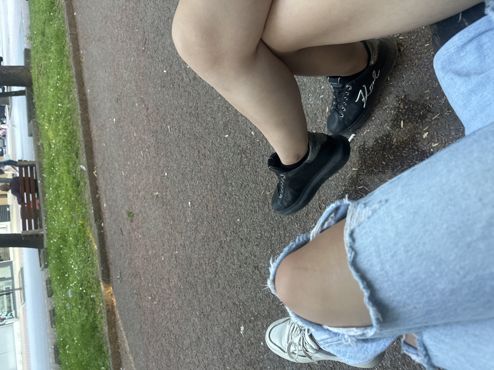 The image captures a close-up view of two individuals' legs and feet as they sit on a bench in a park. Both are wearing casual attire, with one person sporting ripped light-wash jeans and white sneakers, and the other wearing dark sneakers and light-colored shorts or a skirt. The ground beneath them is paved with asphalt, and a strip of green grass with small white flowers borders the path. In the background, a wooden bench is visible, occupied by a person, and further back, a street with passing cars and buildings suggests an urban setting. The scene appears to be in Shumen, Bulgaria, as indicated by the prompt.