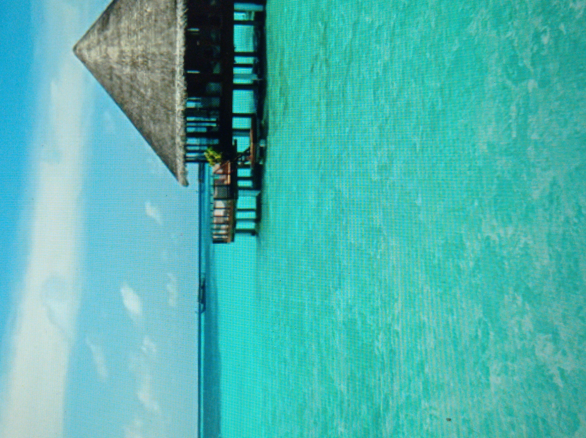 The image depicts an overwater bungalow with a thatched roof, supported by stilts, situated in a vibrant turquoise body of water. The sky above is a bright blue with scattered white clouds. In the distance, on the horizon, a small boat can be seen. The foreground is dominated by the shimmering water, reflecting the light and creating a serene, tropical atmosphere. The bungalow itself appears to be constructed of dark wood and has open-air sections revealing glimpses of interior structures. There are no people visible in the image. The scene suggests a resort or vacation destination. This image does not depict the city of Mukachevo, Ukraine.