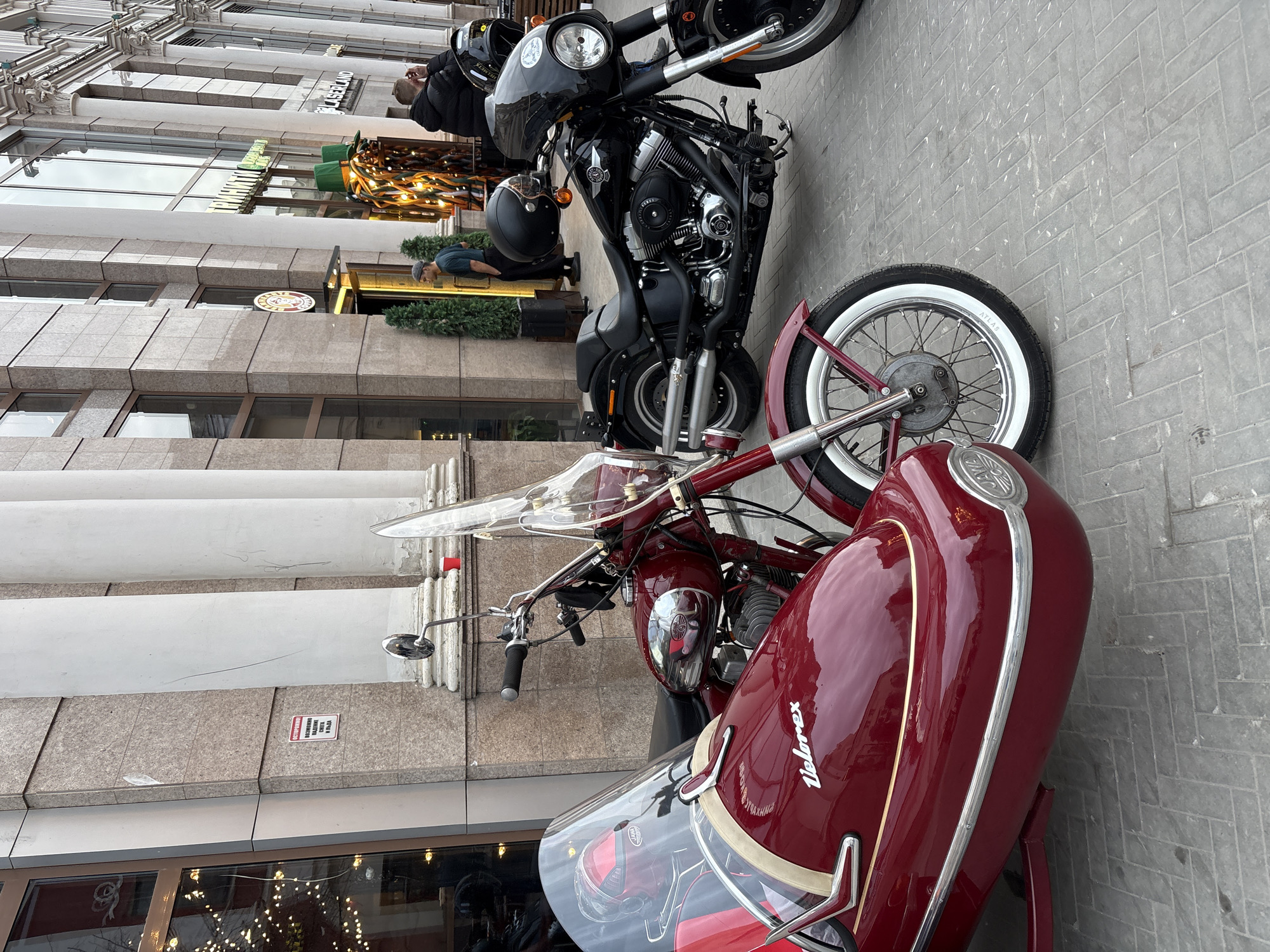 Two motorcycles are prominently displayed on a paved sidewalk in front of a multi-story building. The image is set in an urban environment, likely Kazan, Russia, as indicated by the context.
The foreground features a vintage-style, glossy deep red motorcycle with a matching sidecar. The sidecar bears the name "Velorex" in white script and has a clear windshield and chrome accents. The fuel tank of this motorcycle has "JAWA" visible. Its front wheel features a white sidewall tire with "ATLAS" written on it.
Behind the red motorcycle is a larger, black cruiser-style motorcycle, possibly a Harley-Davidson given its design and visible engine details. A black helmet hangs from its handlebars.
Several people are visible in the background. One person in a dark jacket and cap is near the black motorcycle, while another in a blue shirt and cap stands further back near a shop entrance.
The surrounding buildings are multi-story, with a mix of modern and classical architectural elements, including large windows, white pillars, and stone cladding. Decorative green shrubs are placed outside some establishments. Reflections of the sky and surroundings are visible in the building windows. The ground is paved with light-colored, herringbone-patterned bricks.
Visible text on the buildings includes "LASERLAND" on an upper facade, and a red warning sign lower down that reads "ВНИМАНИЕ СОСУЛЬКИ С КРЫШИ" (ATTENTION ICICLES FROM THE ROOF). The lighting suggests daytime, possibly overcast. The motorcycles appear to be parked, and the people are casually present in the urban scene.