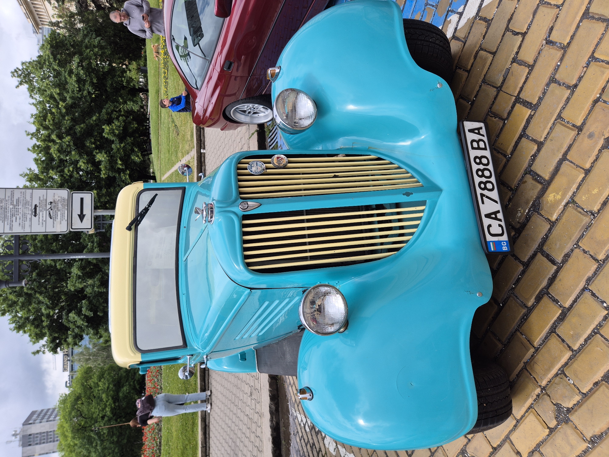 The image shows a turquoise vintage car parked on a cobblestone street. The car's license plate reads "CA 7888 BA," indicating it is registered in Sofia, Bulgaria. A second, red car is partially visible behind the turquoise car. In the background, there is a grassy area with trees and a few people. One person is taking a photo with a selfie stick. The Grand Hotel Millennium Sofia is visible in the background, suggesting the location is near the city center. A street sign is partially visible, indicating parking regulations. A man is visible behind the red car with a dog.