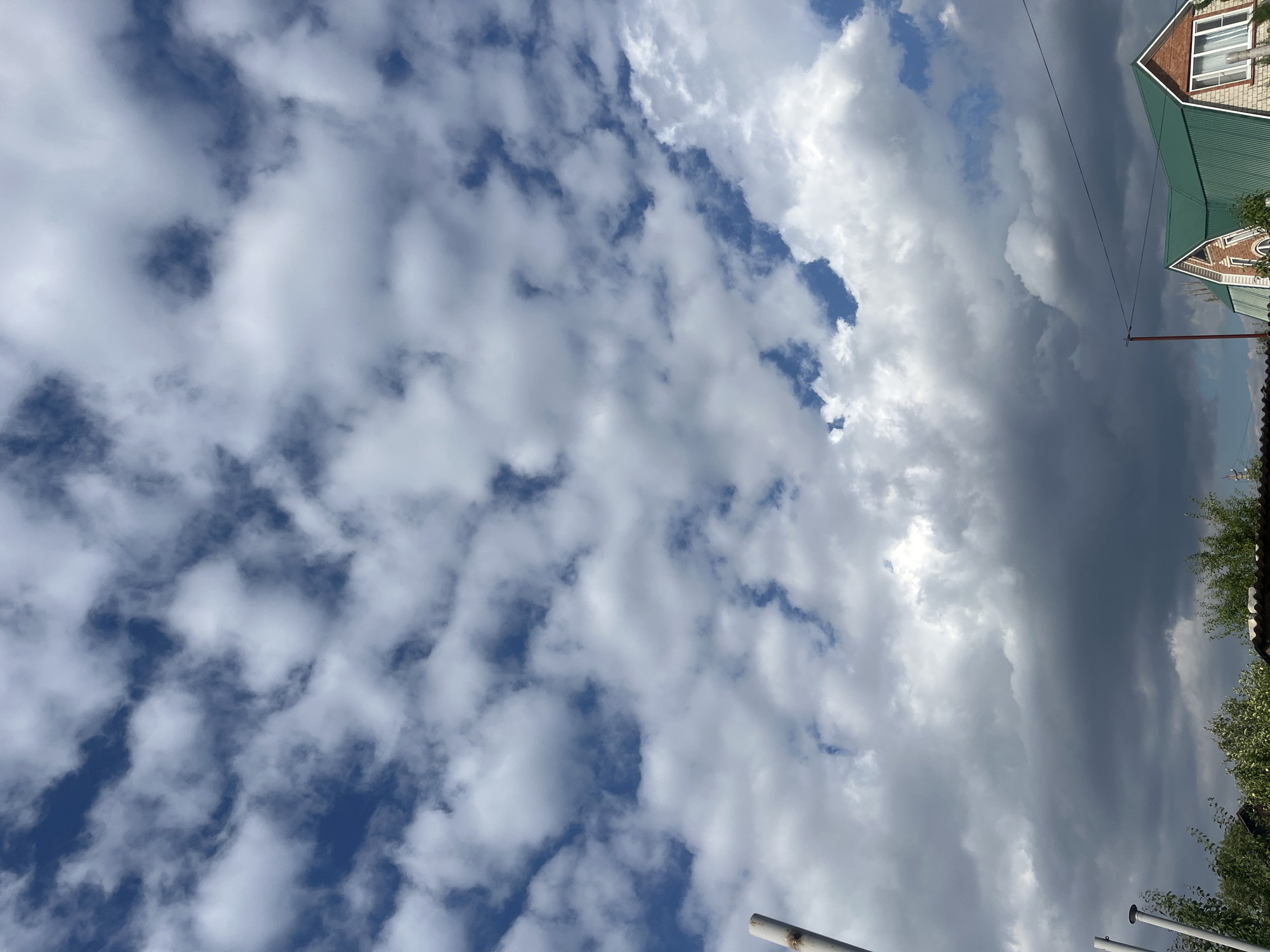 The image displays a cloudy sky above a residential area. The clouds are a mix of white and dark gray, suggesting a dynamic weather pattern. In the lower portion of the frame, a building with a green roof and brick facade is visible. Trees and other structures are also present, indicating a suburban or rural setting. The scene appears to be captured from a low angle, emphasizing the sky. The location is identified as Tikhoretsk, Russia.