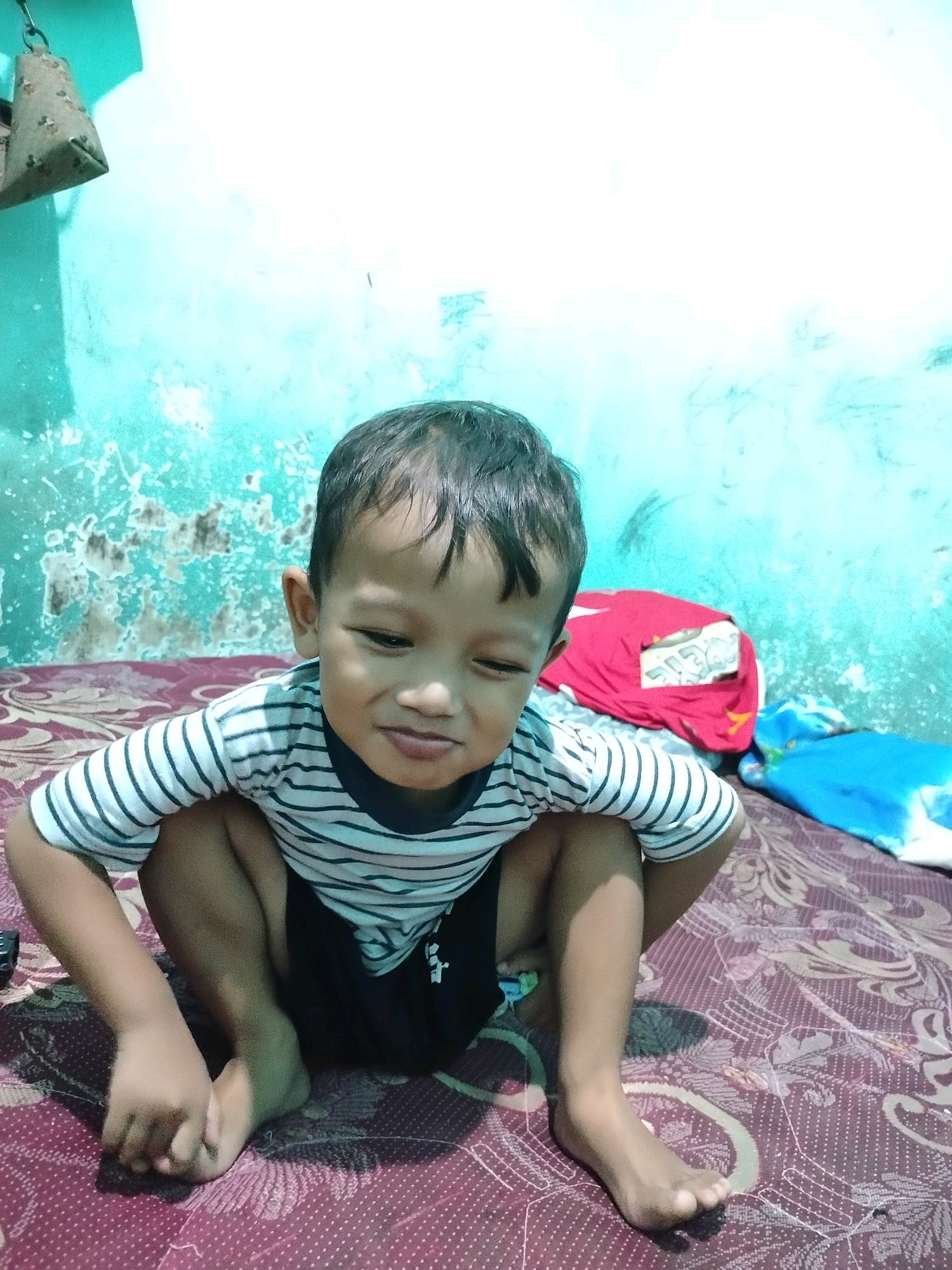 A young boy of Southeast Asian descent, appearing to be between 3-5 years old, is photographed sitting on a patterned bed, likely in a domestic setting in Beloh, Indonesia. He is wearing a striped t-shirt with black shorts. His body is in a squatting position, with his knees bent and his arms resting on his legs. His facial expression is neutral, with his eyes partially closed. The background shows a teal-colored wall with a distressed texture. To the right of the boy, there are two folded blankets, one red and one blue, on the bed. A bell-shaped object is suspended from the ceiling in the upper left corner of the image. The lighting suggests an indoor environment with artificial light.