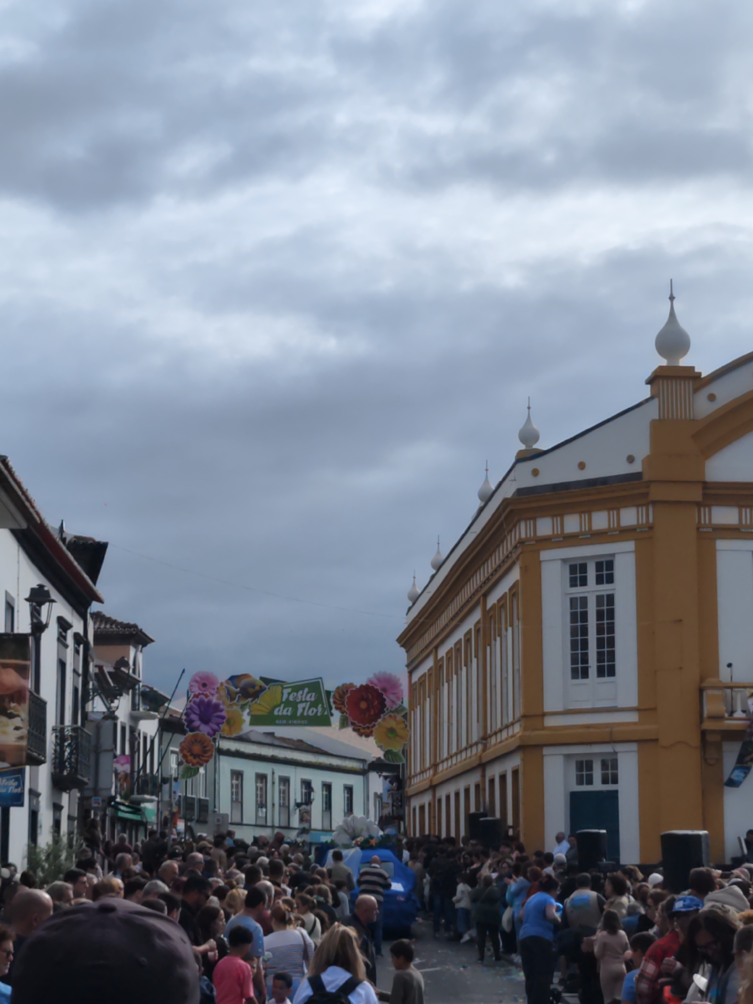 The image depicts a street scene in Ribeira Grande, Portugal, during the "Festa da Flor" flower festival. A large crowd of people fills the street, suggesting a public celebration or parade. Overhead, flower decorations are strung across the street, including a banner that reads "Festa da Flor". The buildings in the background are a mix of white and brown, with some of them featuring traditional Portuguese architectural details. The sky is overcast, with heavy gray clouds. Speakers are placed on the side of the road near a white and brown building.
