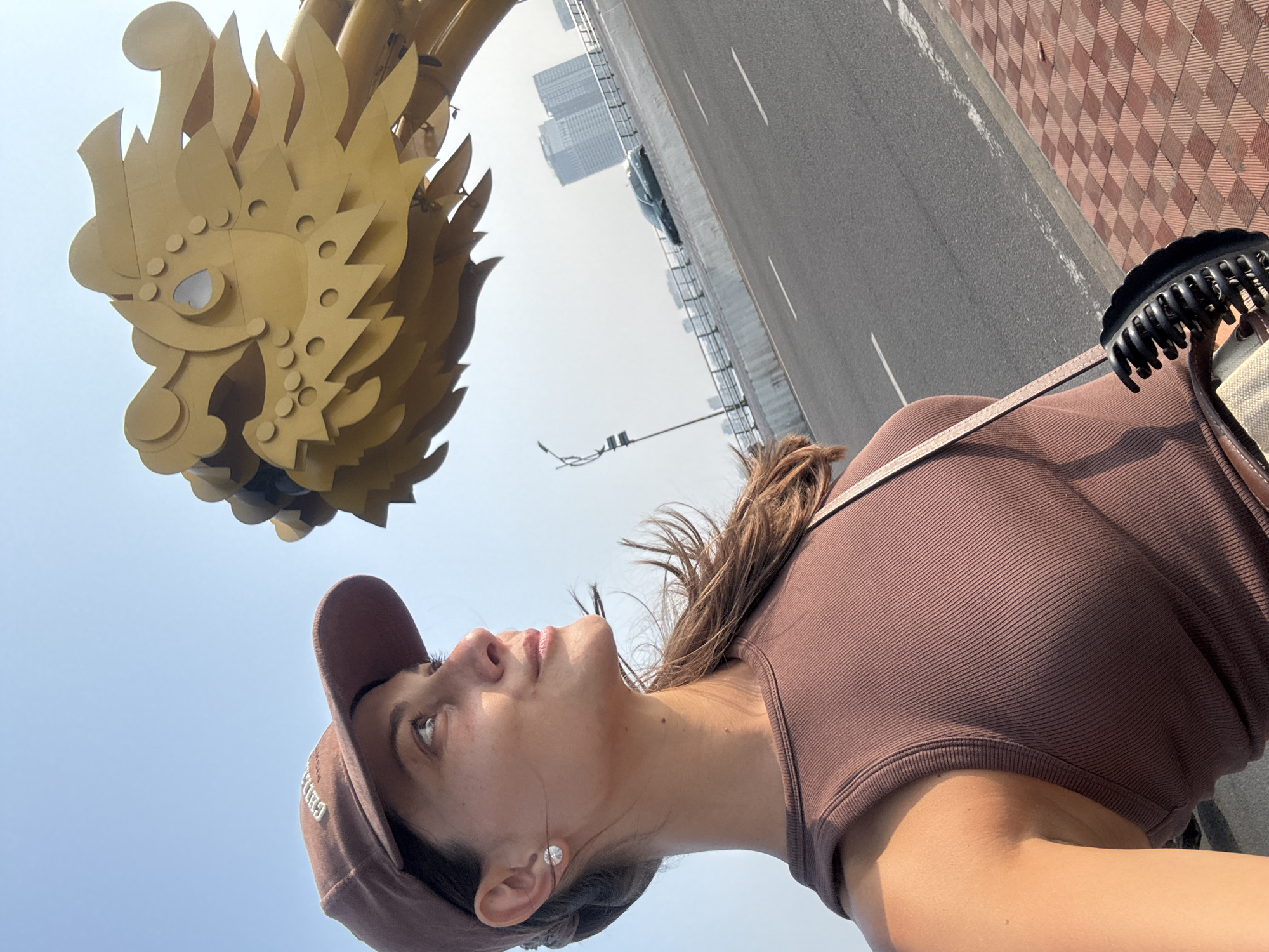 A young woman is gazing up at the head of a large golden dragon sculpture. She is wearing a brown baseball cap and a brown tank top. A section of roadway is visible in the background, with some buildings in the distance. The dragon is located on Dragon Bridge in Đà Nẵng, Vietnam.