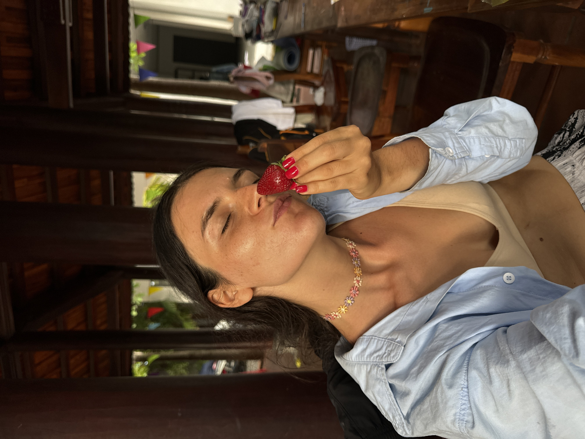 A woman is lying down, holding a strawberry up to her lips. Her eyes are closed and she appears to be enjoying the moment. She is wearing a light blue button-down shirt over a beige tank top and a colorful floral choker necklace. Her fingernails are painted red. The background suggests an outdoor setting with wooden structures and some greenery visible. The image is taken from a close-up perspective, focusing on the woman and the strawberry. The lighting suggests it might be daytime.