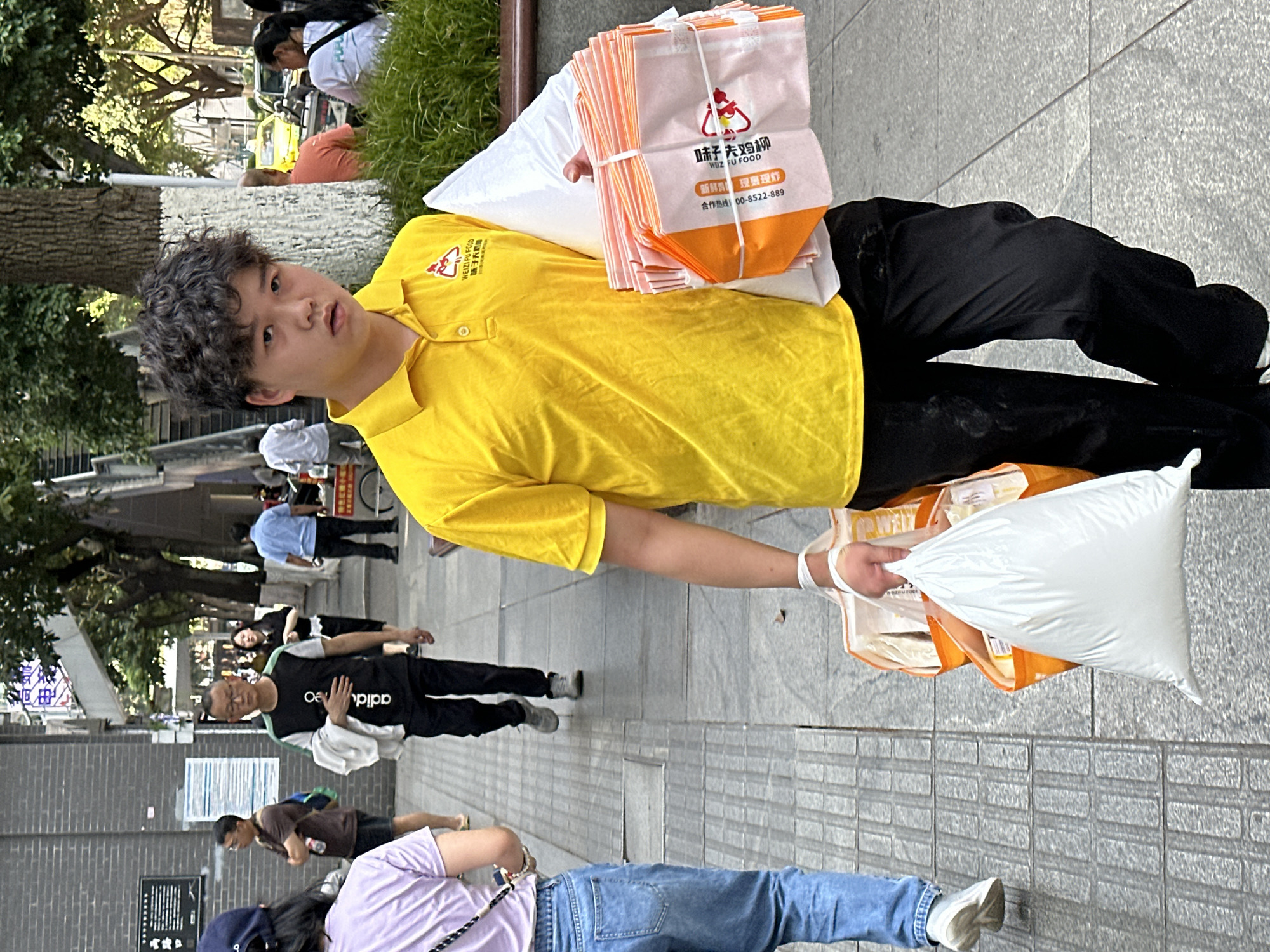 A young person is standing on a city sidewalk in Yuzhong District, China. The individual is wearing a bright yellow polo shirt and black pants. They are holding several orange and white delivery bags, with the logo "WEIZI FU FOOD" and Chinese characters visible on them. In the background, other pedestrians can be seen walking on the sidewalk. The street is bordered by buildings and trees, creating an urban environment.