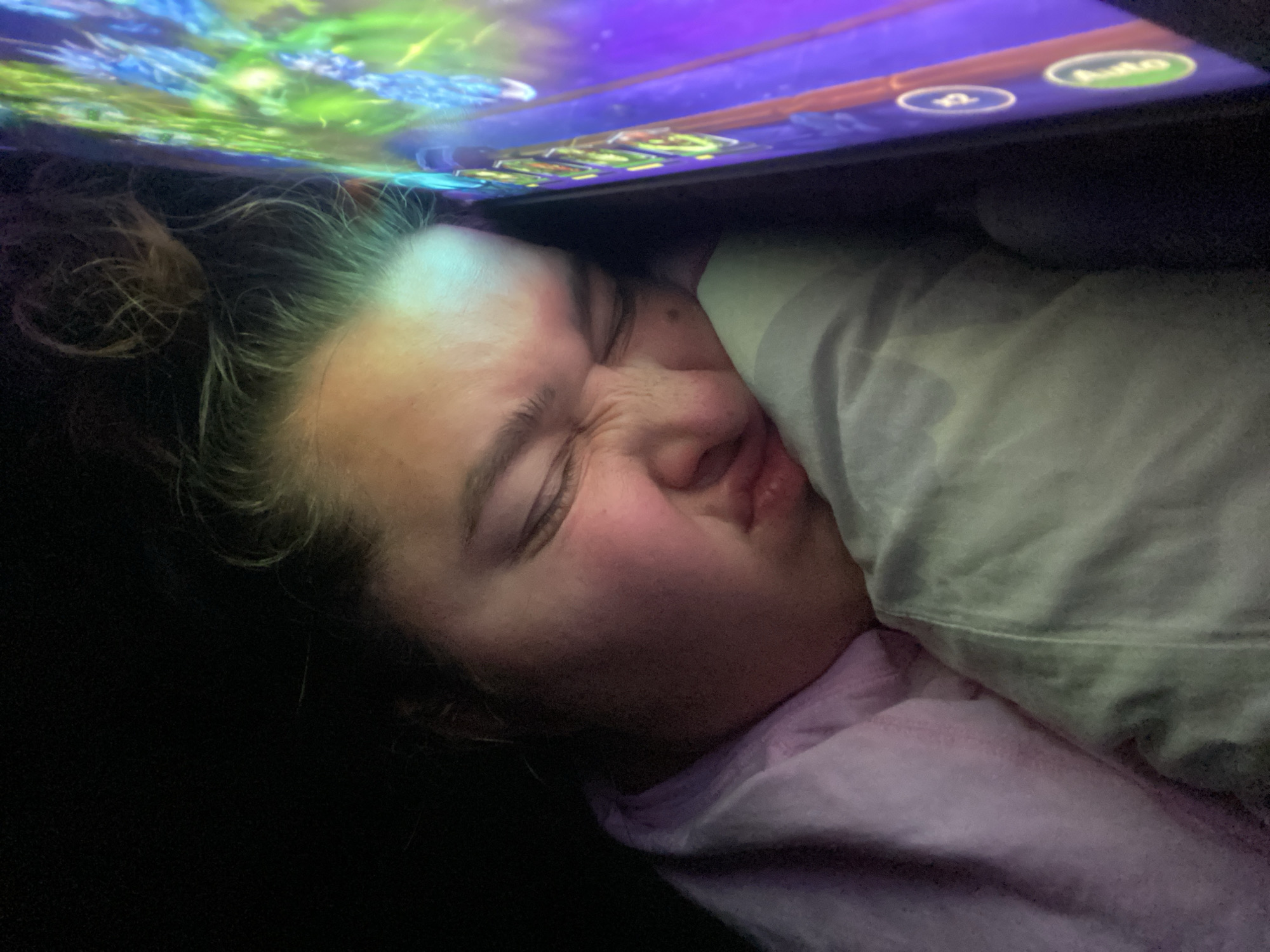 The image shows a person, likely a young female, sleeping with her face pressed against a pillow. Her eyes are closed, and her mouth is slightly open. She is wearing a pink hooded garment. A tablet device is positioned in front of her, reflecting light onto her face. The screen of the tablet displays a colorful, vibrant interface with what appear to be game elements. The background is dark, suggesting a dimly lit environment, possibly nighttime. The scene is captured at a close-up, personal angle. The provided context places this image in the city of Perm, Russia.